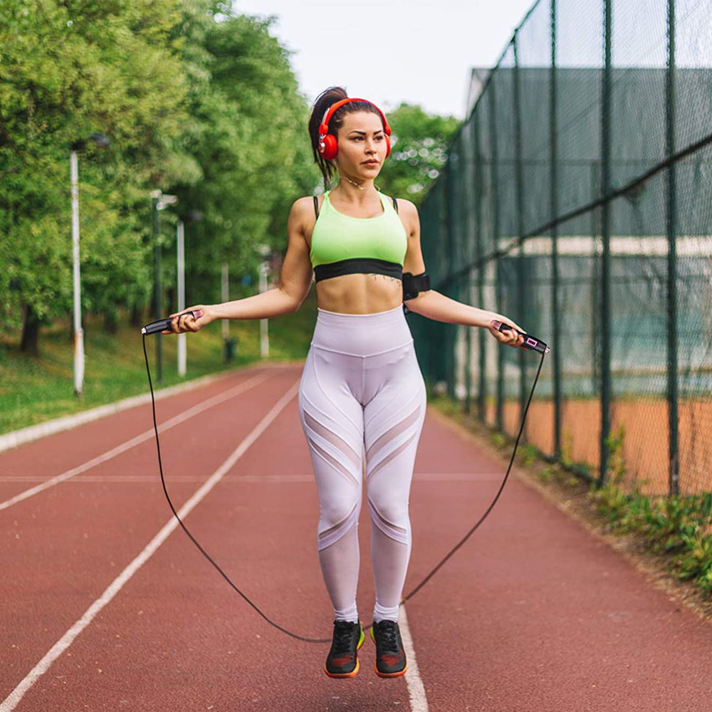 Eine Frau springt auf einer Bahn Seil. Sie trägt einen grünen Sport-BH, weiße Leggings und rote Kopfhörer. Im Hintergrund sind ein Zaun und Bäume zu sehen.