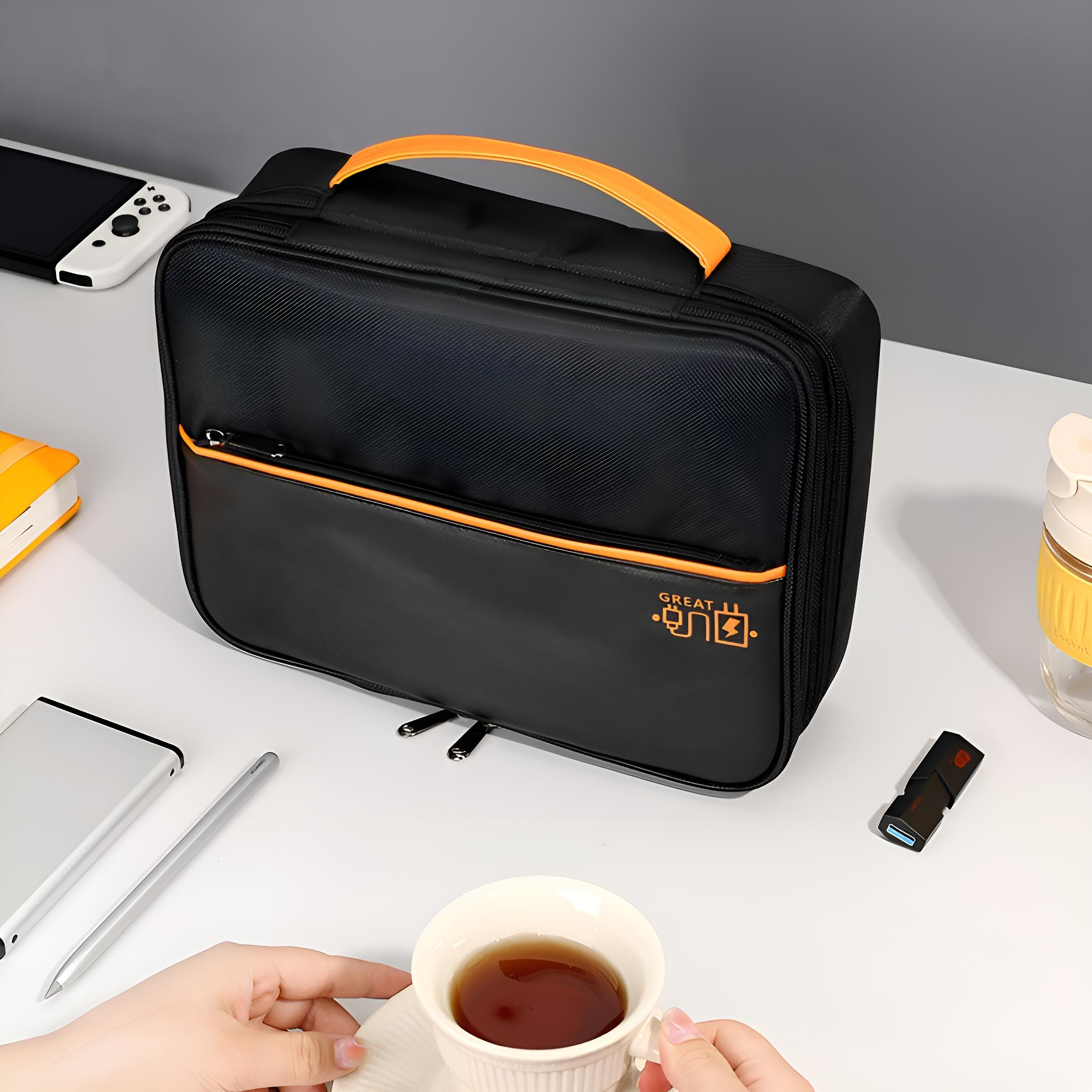 Schwarze Tasche mit orangefarbenem Griff auf weißem Schreibtisch. Teetasse, Stift, USB-Stick und Switch sichtbar. Hände halten Tasse.