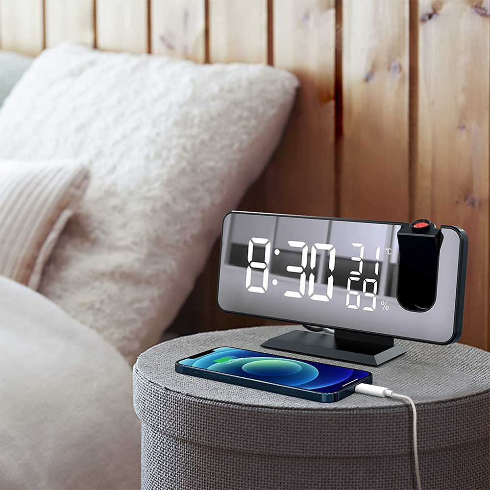 Eine Digitaluhr zeigt 8:30 Uhr an. Ein Telefon wird auf einem Hocker aufgeladen. Im Hintergrund Kissen und ein hölzernes Kopfteil.