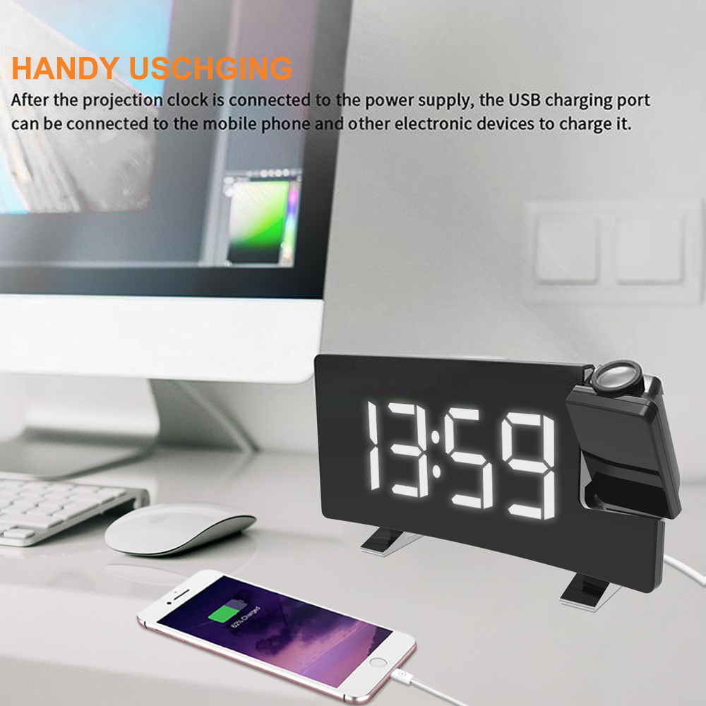 Eine Digitaluhr mit der Anzeige 13:59, ein ladendes Telefon und ein Computermonitor stehen auf einem Schreibtisch. Die Uhr ist schwarz und die Zahlen weiß.