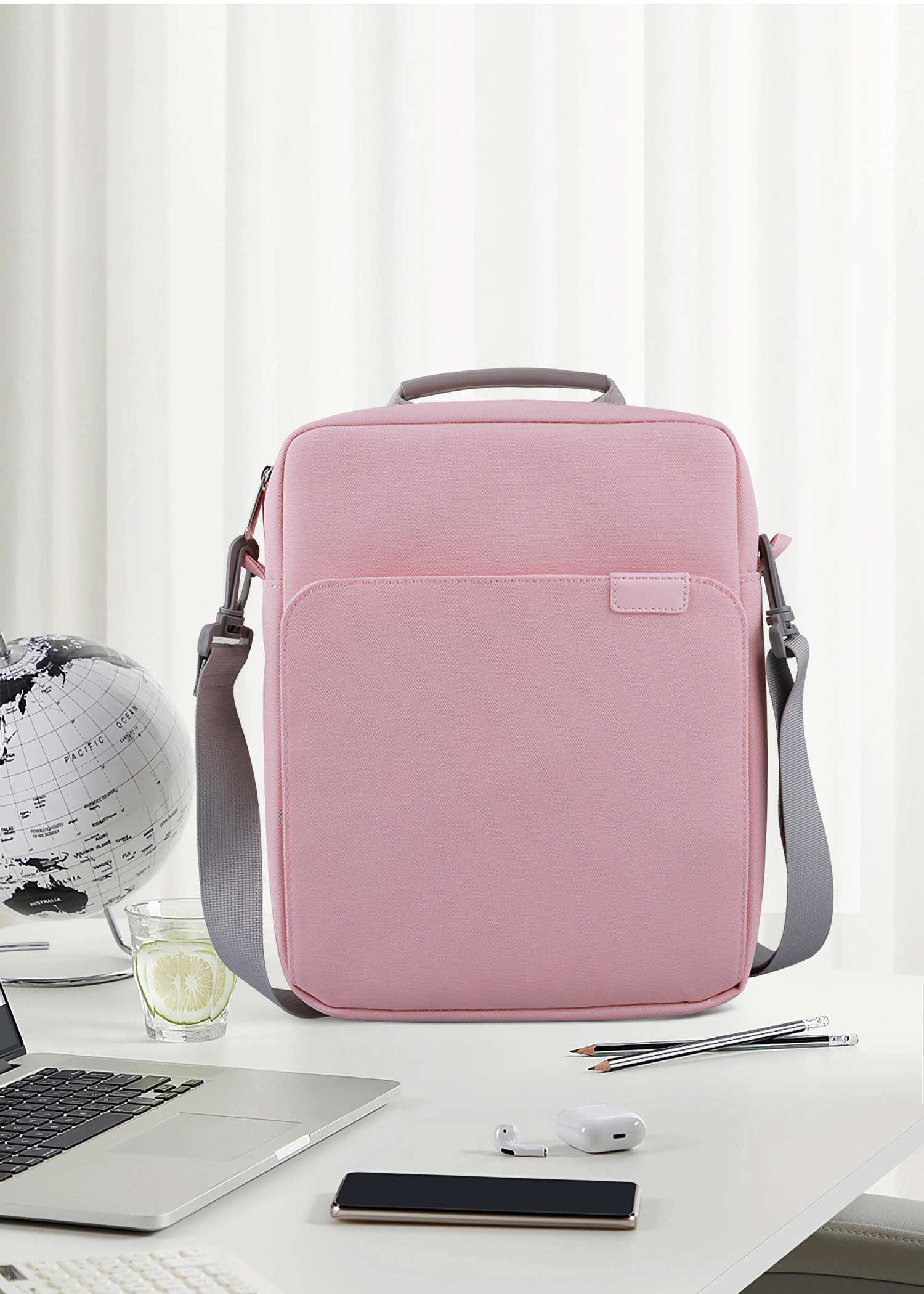 Eine rosa Umhängetasche mit grauem Gurt liegt auf einem weißen Schreibtisch mit Laptop, Globus, Wasserglas und Bleistiften.