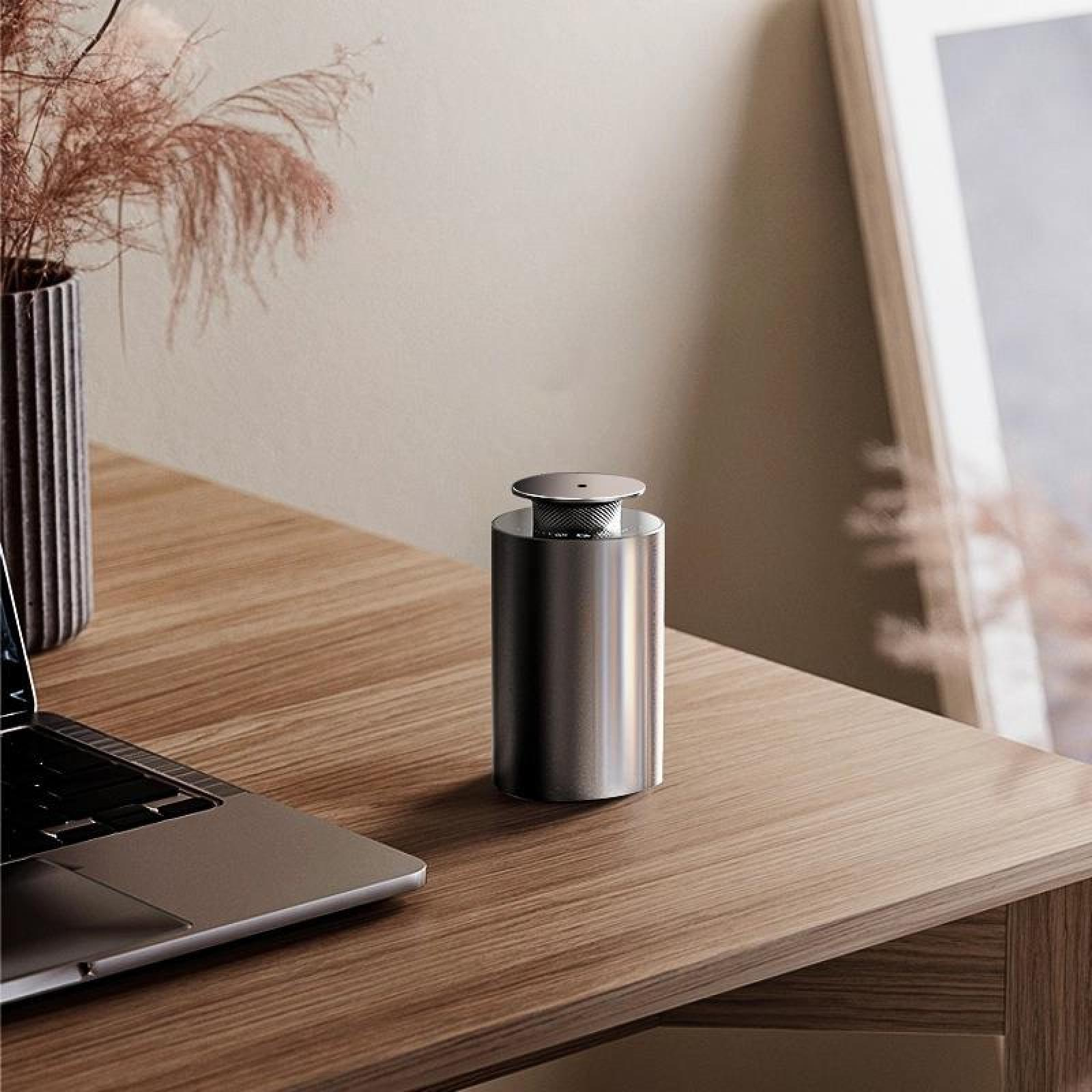 Ein silberner zylindrischer Gegenstand steht auf einem Holztisch neben einem Laptop und einer Vase mit getrockneten Pflanzen. Im Hintergrund ein Bilderrahmen.