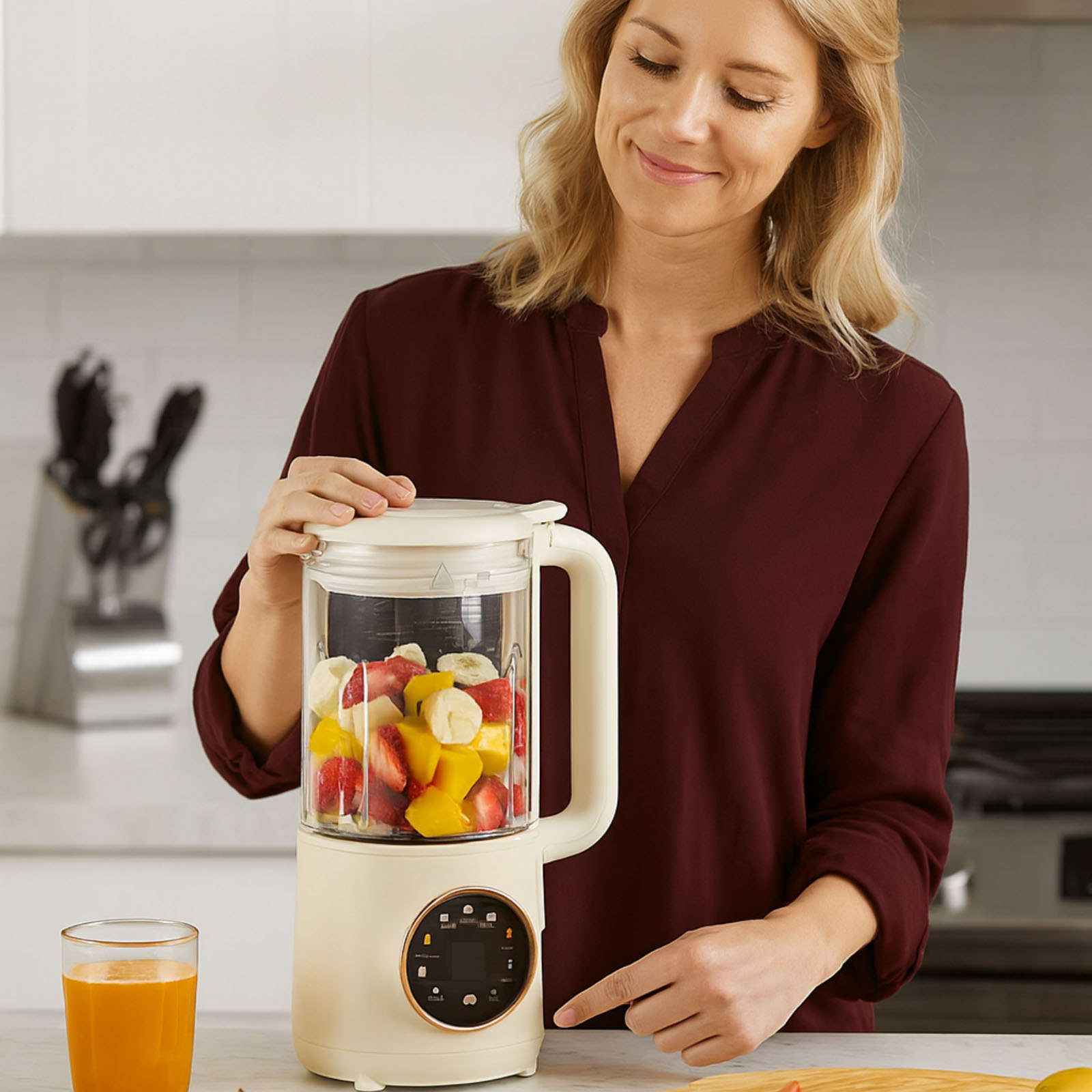 Eine Frau mit blonden Haaren lächelt einen Mixer an, der mit Früchten gefüllt ist. Daneben steht ein Glas Saft. Sie ist in einer Küche.