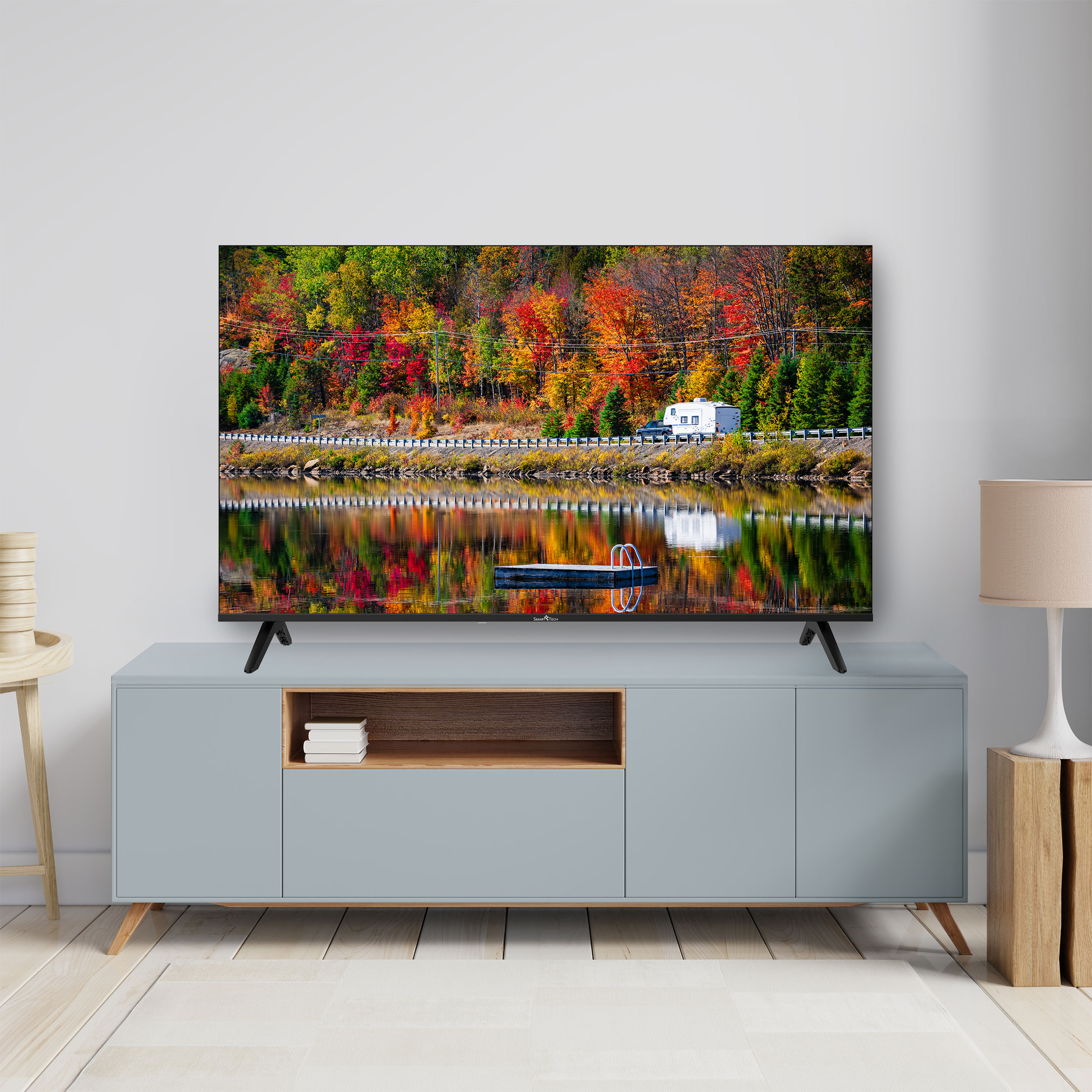 Una TV mostra un paesaggio autunnale colorato. La TV è su un mobile blu. Una lampada e uno sgabello sono ai lati.