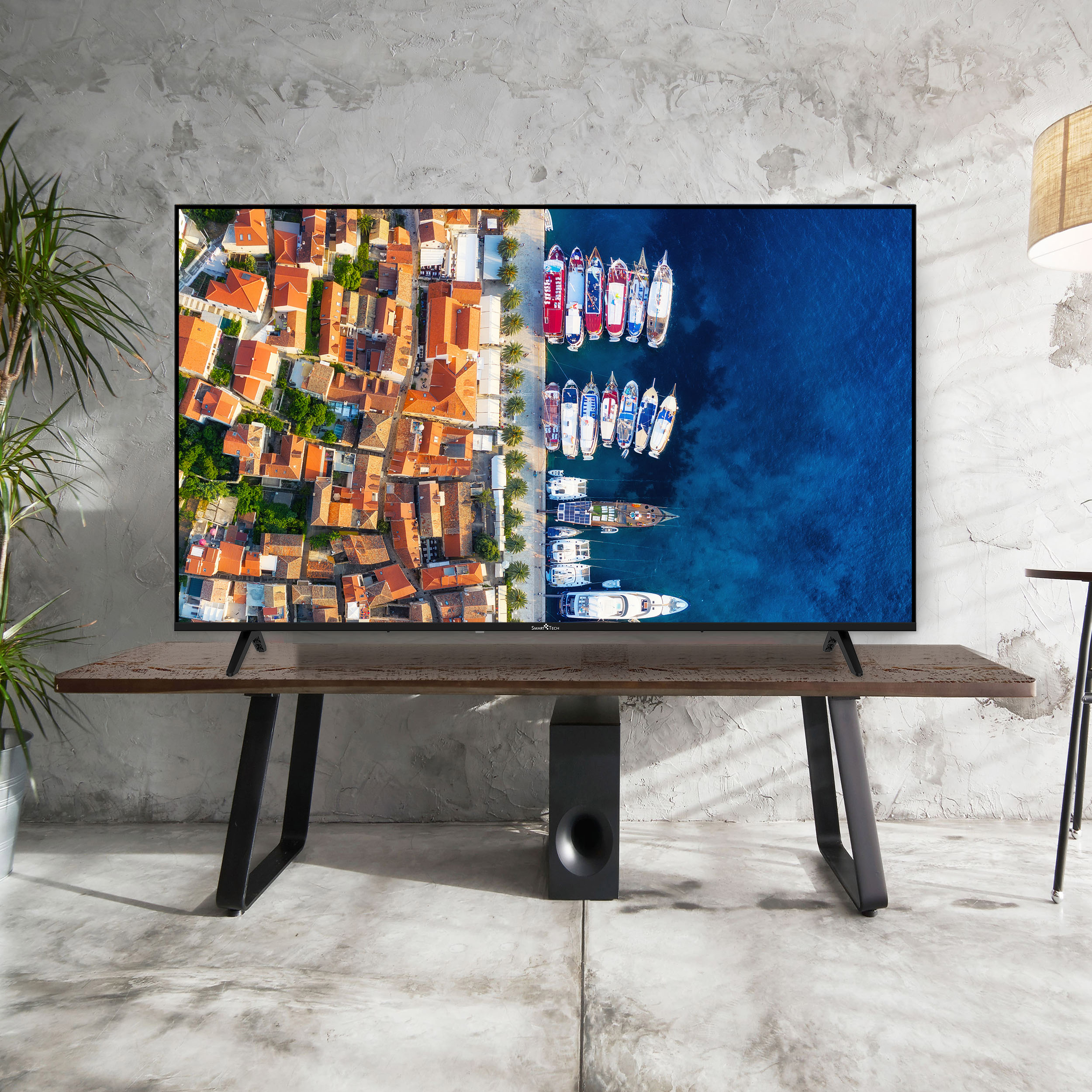 Fernseher auf einem Tisch zeigt eine Küstenstadt und Boote. Graue Wand dahinter. Pflanze, Lampe und Subwoofer sichtbar.