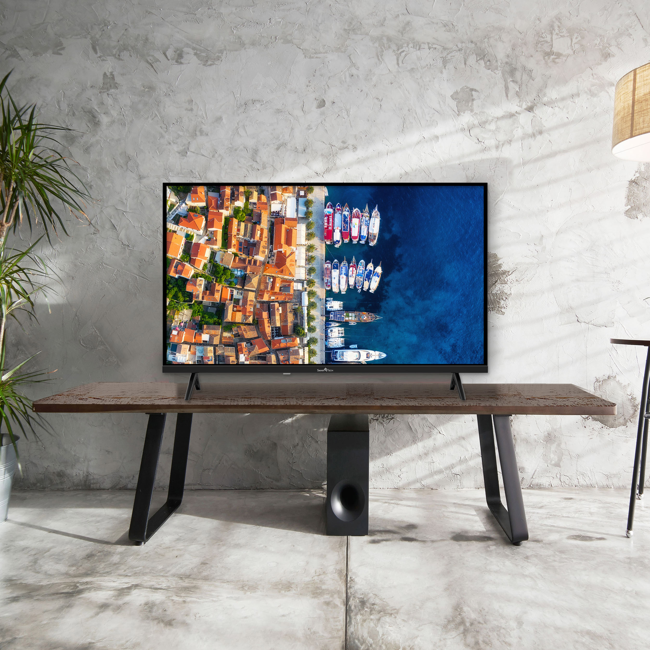 Un televisor muestra un pueblo costero. El televisor está sobre una mesa de madera con patas negras. Debajo hay un altavoz.