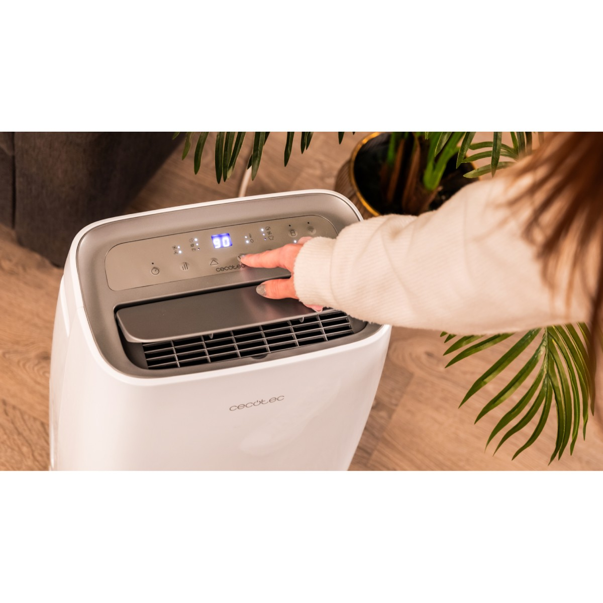 Dłoń kobiety na białym oczyszczaczu powietrza. Ma cyfrowy wyświetlacz z temperaturą. Zielone rośliny w tle.
