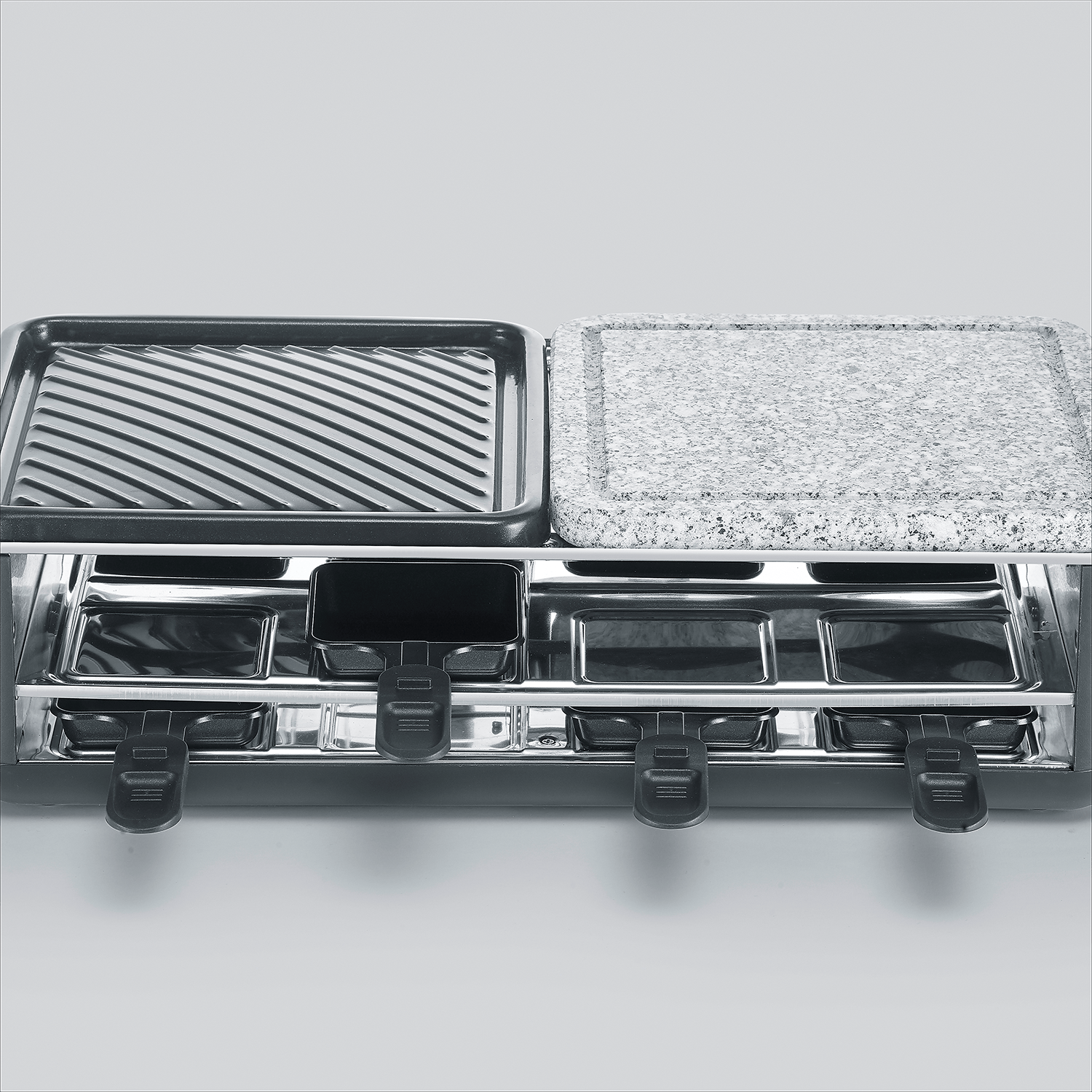 Grill do raclette z żebrowaną płytą grillową i kamienną płytą grillową. Poniżej małe tacki z uchwytami.