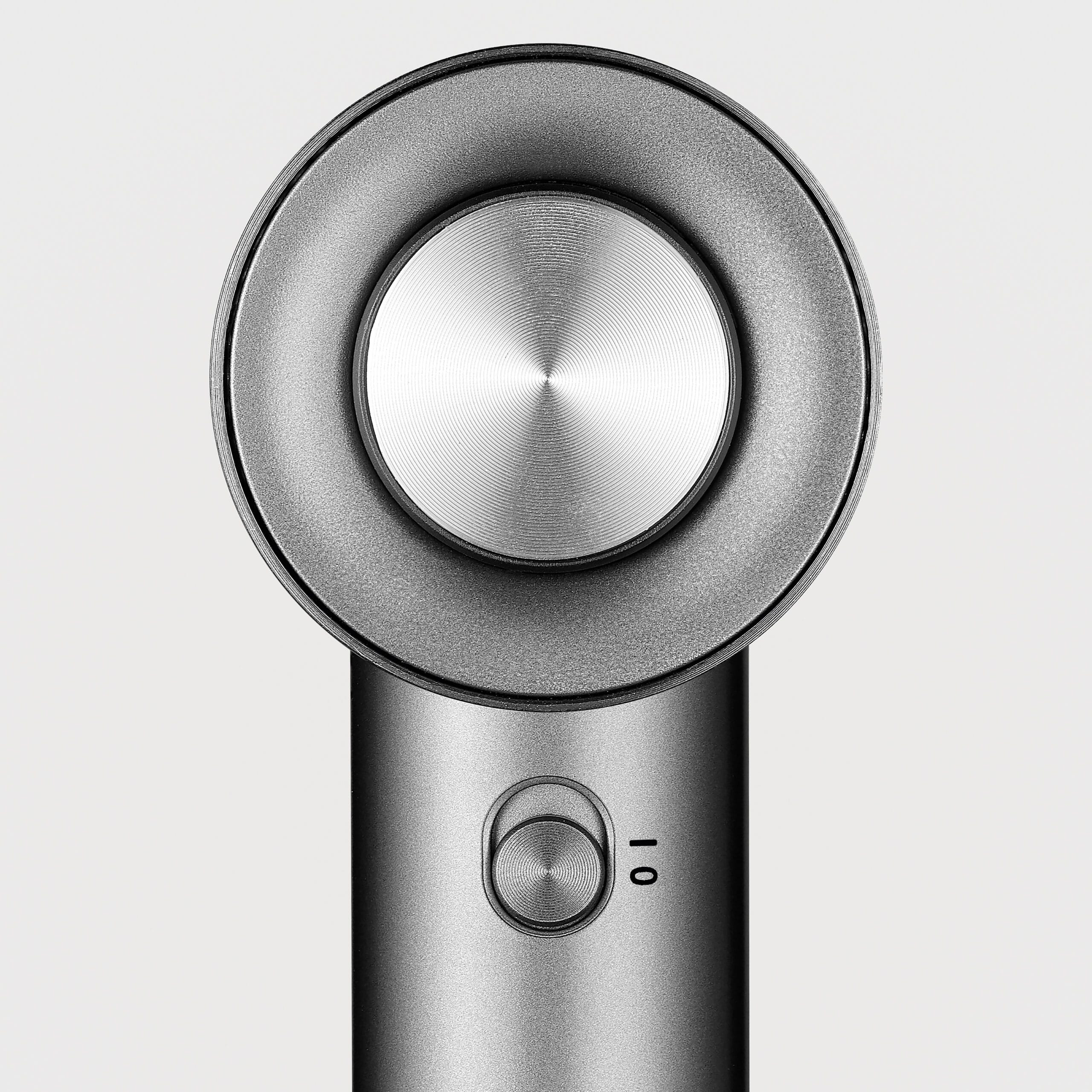 Zbliżenie srebrnej suszarki do włosów. Ma okrągłą głowicę z centralnym przyciskiem i przyciskiem zasilania poniżej. Tło jest białe.