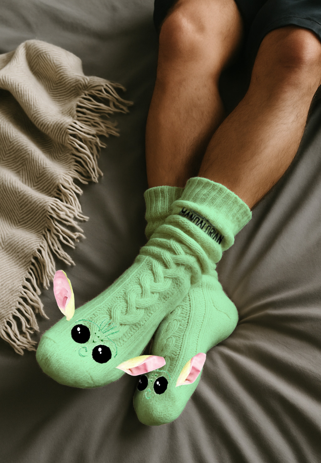 Person trägt grüne Socken, gezeichnet als Baby Yoda. Beine und eine Decke sind ebenfalls sichtbar. Auf den Socken steht 'Mandalorian'.