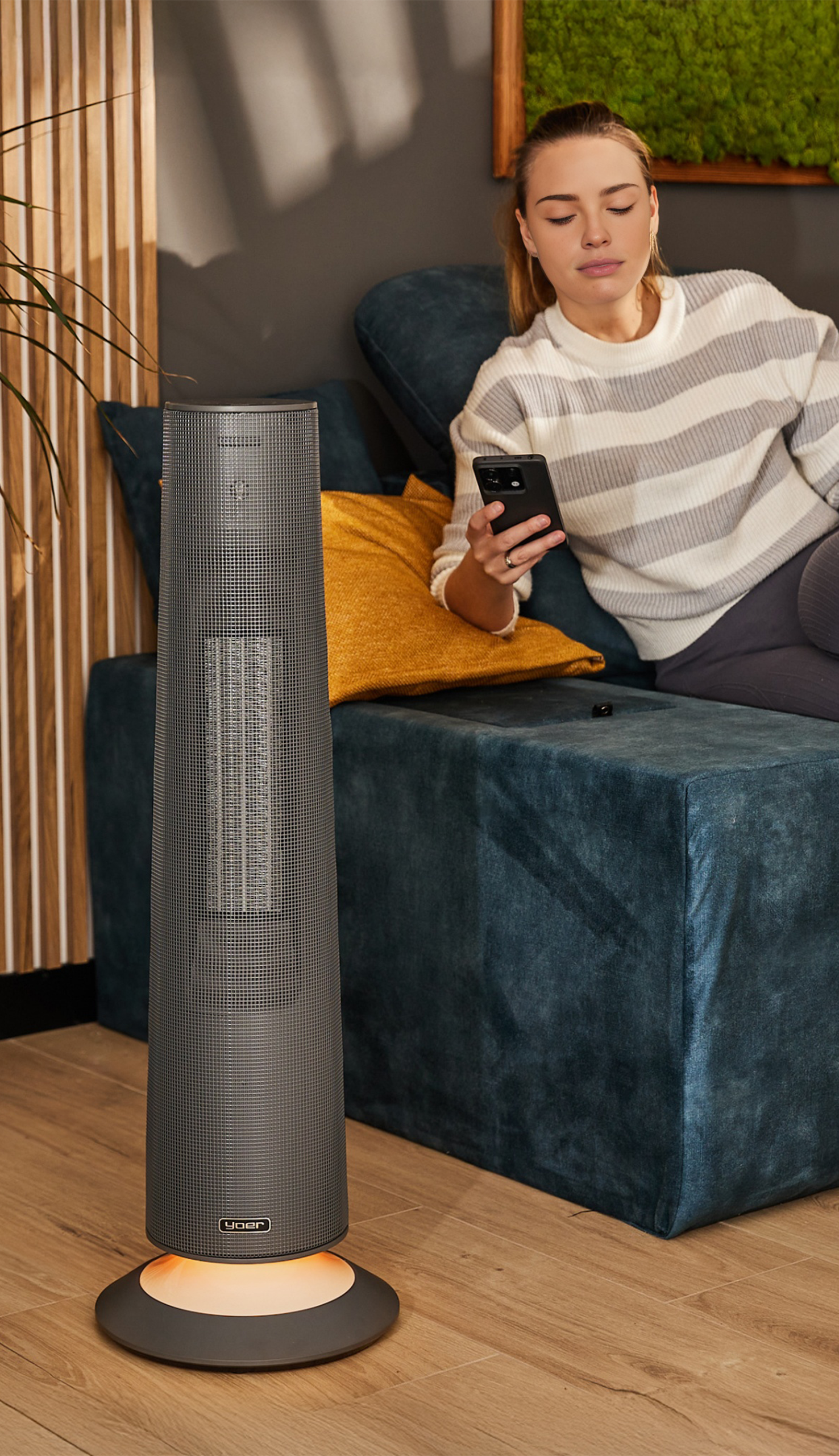 Kobieta siedzi na turkusowej sofie trzymając telefon. Szary grzejnik stoi z przodu. Tło ma drewniane listwy i dzieło sztuki z mchu.