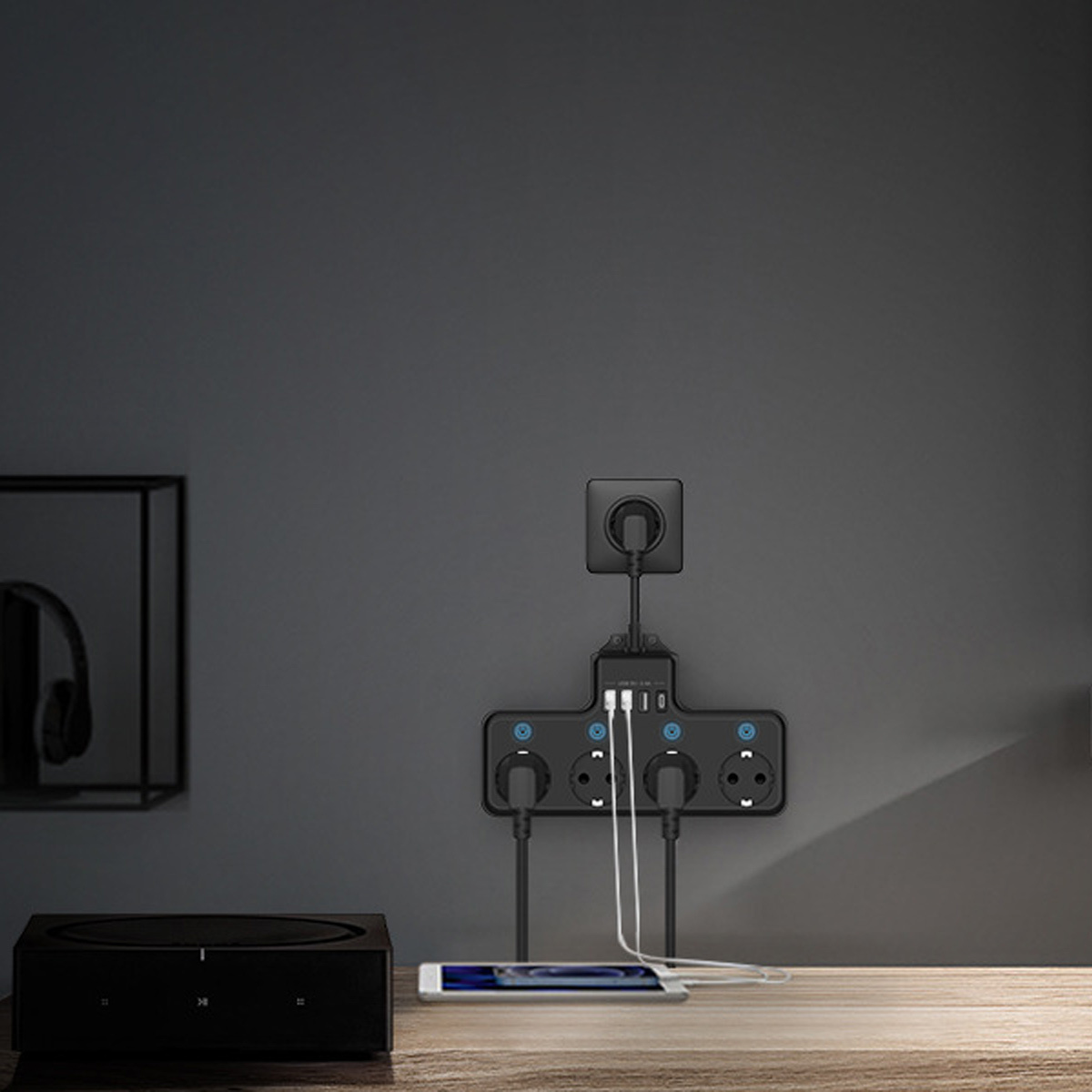 Schwarze Steckdosenleiste mit Steckdosen und USB-Anschlüssen, Telefon lädt, an der Wand befestigt. Lautsprecher und Kopfhörer im Hintergrund.