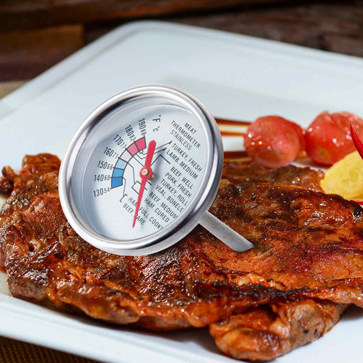 Ein Fleischthermometer in einem Steak zeigt die Temperatur an. Tomaten und ein gelbes Element liegen neben dem Steak auf einem weißen Teller.