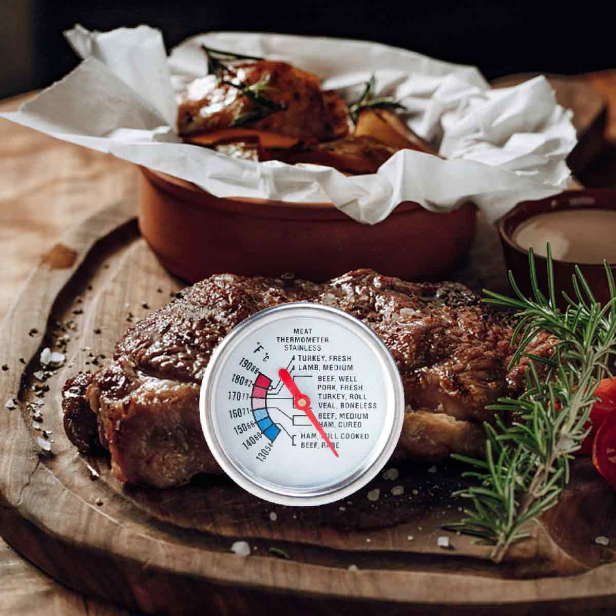 Ein gebratenes Steak mit einem Fleischthermometer auf einem Holzbrett. Daneben Rosmarin und eine Soße. Im Hintergrund gekochtes Essen.
