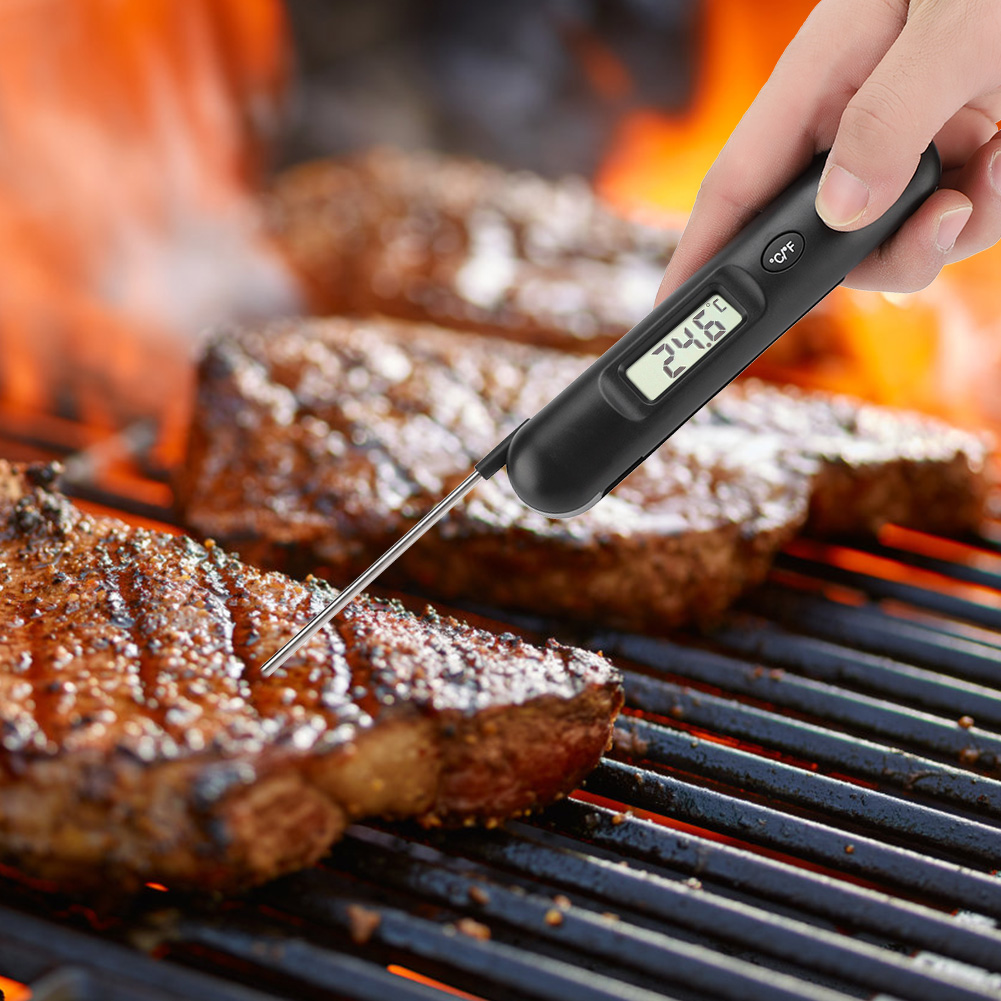 Eine Hand hält ein Thermometer, das die Temperatur von gegrillten Steaks misst. Die Steaks liegen auf einem Grill, im Hintergrund ist ein Feuer.