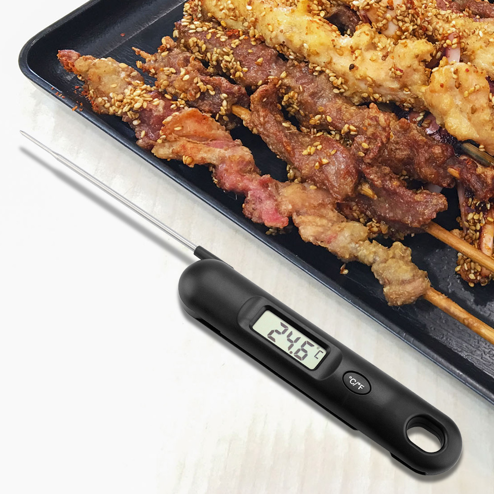 Ein schwarzes digitales Thermometer zeigt 24,6 °C an. Im Hintergrund liegen Spieße mit gegrilltem Fleisch auf einem schwarzen Tablett, einige mit Sesam.