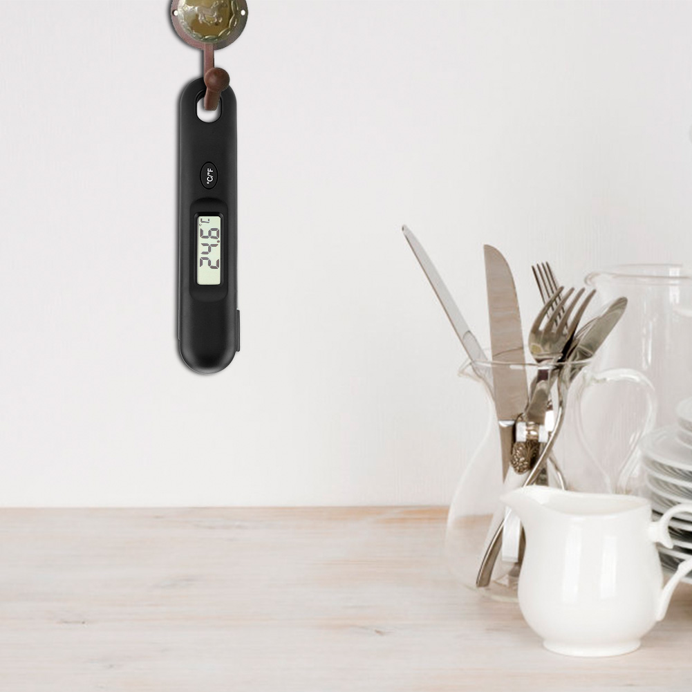 Ein schwarzes digitales Thermometer hängt an einer Wand. Utensilien befinden sich in einer Glasvase und ein kleiner weißer Krug auf einem Holztisch.