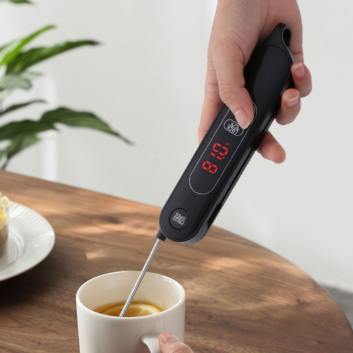 Eine Hand hält ein schwarzes Thermometer in einer weißen Tasse mit Tee. Das Thermometer zeigt 81,0 °F an. Ein Holztisch und eine Pflanze sind im Hintergrund.