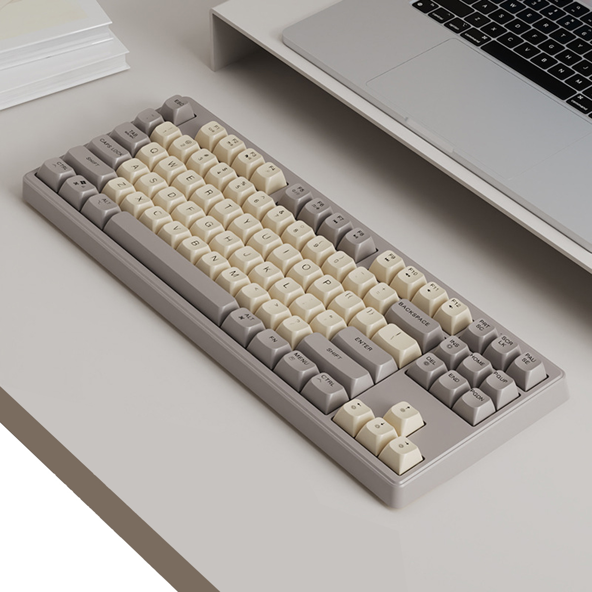 Eine graue und cremefarbene mechanische Tastatur liegt auf einer weißen Oberfläche. Ein Laptop und ein Stapel Bücher sind im Hintergrund zu sehen.