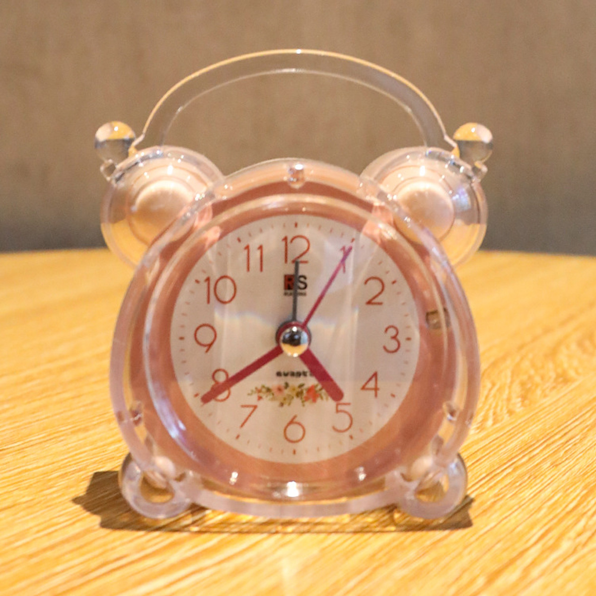 Ein rosa Wecker mit transparentem Gehäuse steht auf einer Holzoberfläche. Die Uhr hat ein weißes Zifferblatt mit roten Zeigern und Zahlen.