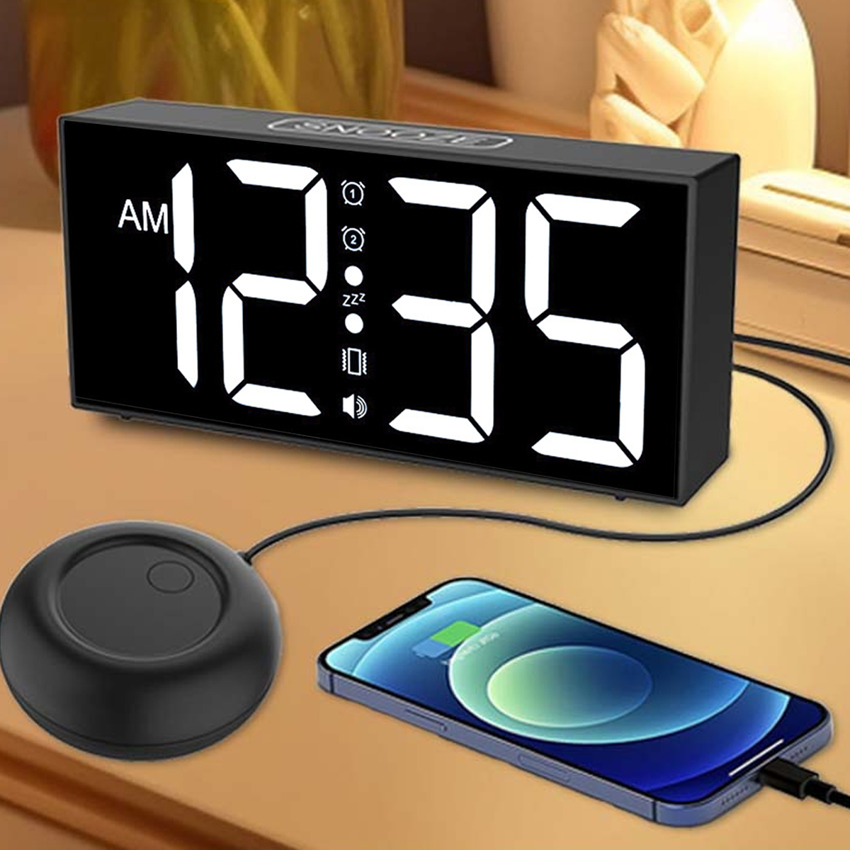 Eine Digitaluhr zeigt 12:35 Uhr an. Daneben lädt ein Telefon. Ein schwarzer Vibrationspuck ist mit der Uhr verbunden.