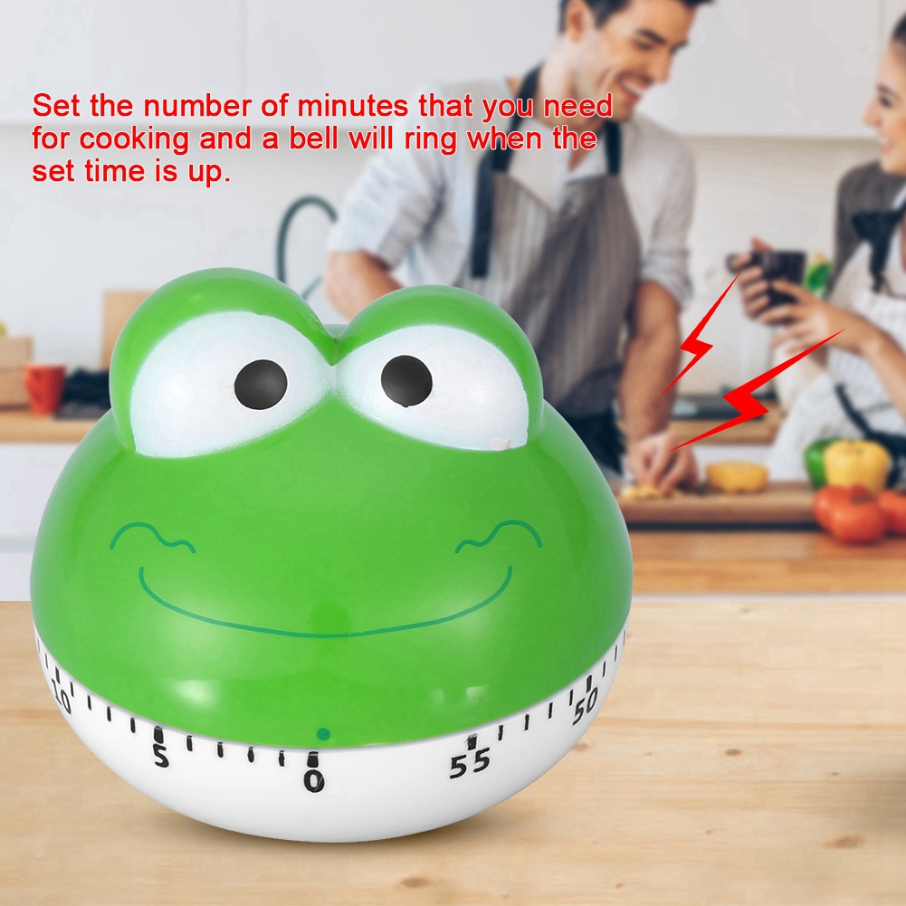 Ein grüner froschförmiger Küchentimer steht auf einer Holzoberfläche. Ein Paar kocht im Hintergrund. Rote Blitze zeigen Ton an.