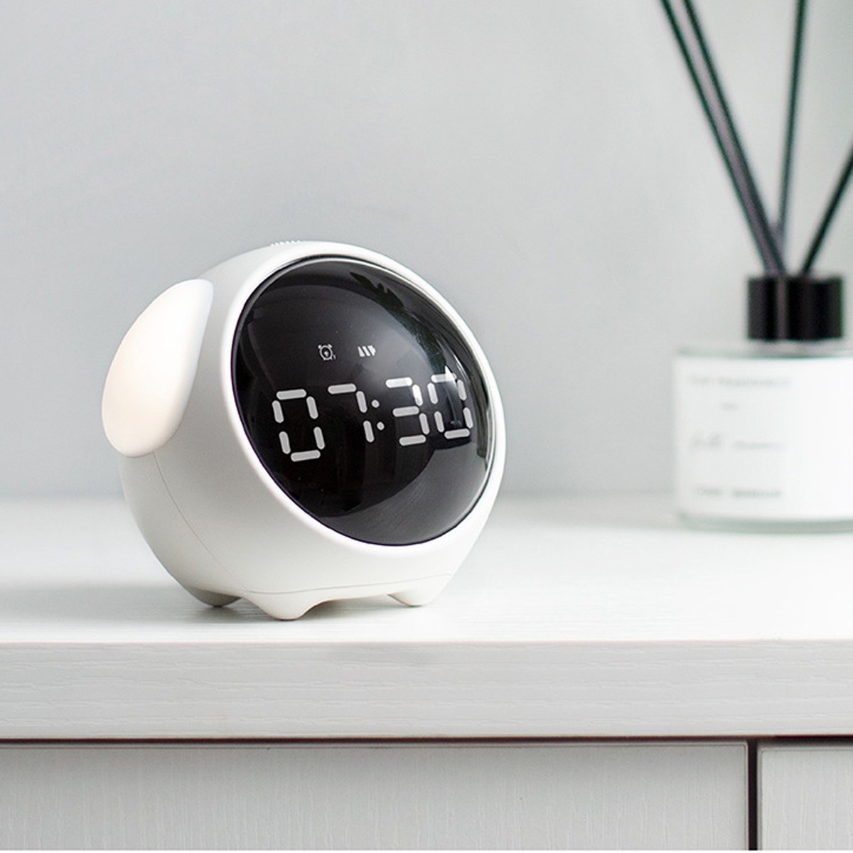 Ein weißer, eiförmiger digitaler Wecker, der 07:30 Uhr anzeigt. Er steht auf einer weißen Oberfläche, im Hintergrund ein Schilfrohrdiffusor.