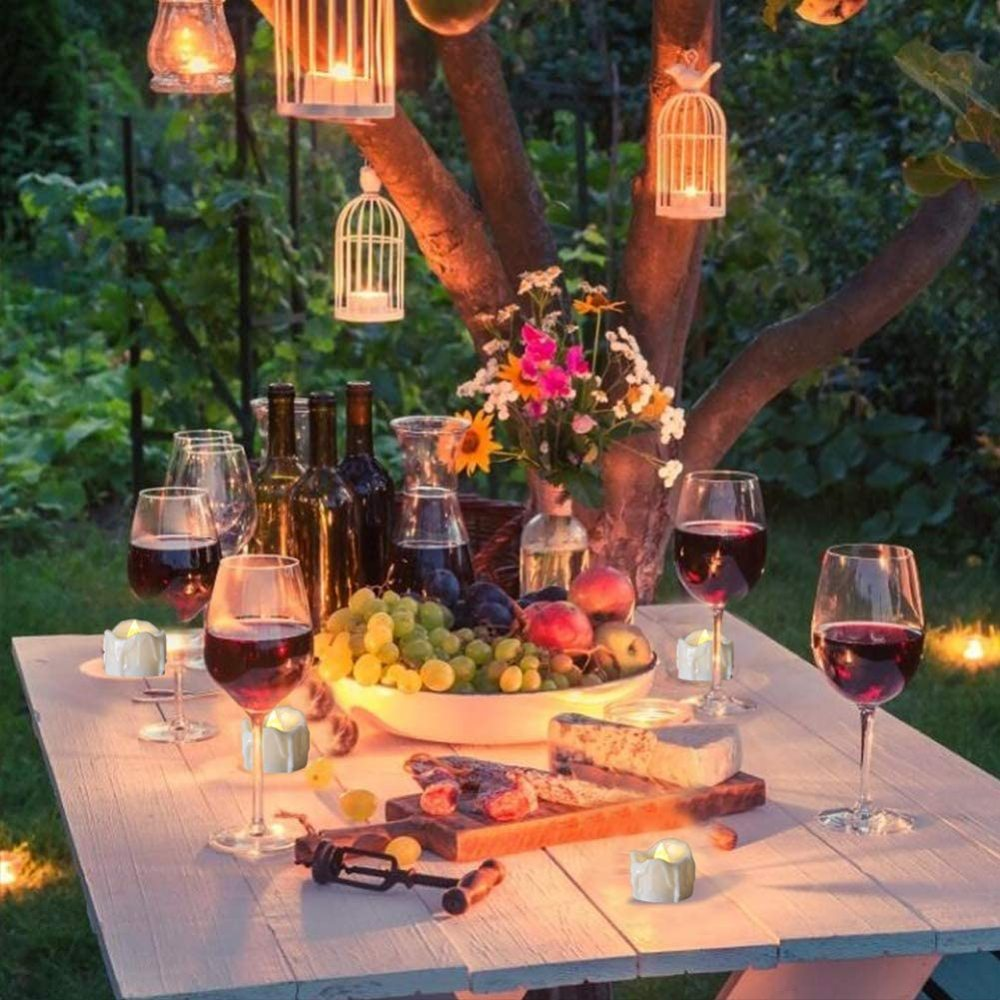 Ein Tisch mit Wein, Obst, Käse und Kerzen unter einem Baum mit hängenden Laternen. Die Szene deutet auf ein Treffen im Freien hin.