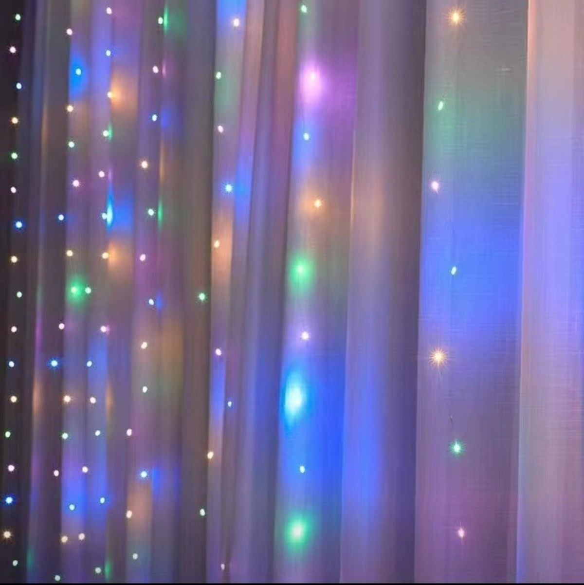 Mehrfarbige Lichterketten hängen vor einem weißen Vorhang. Die Lichter haben ein sanftes Leuchten und schaffen eine festliche Atmosphäre.