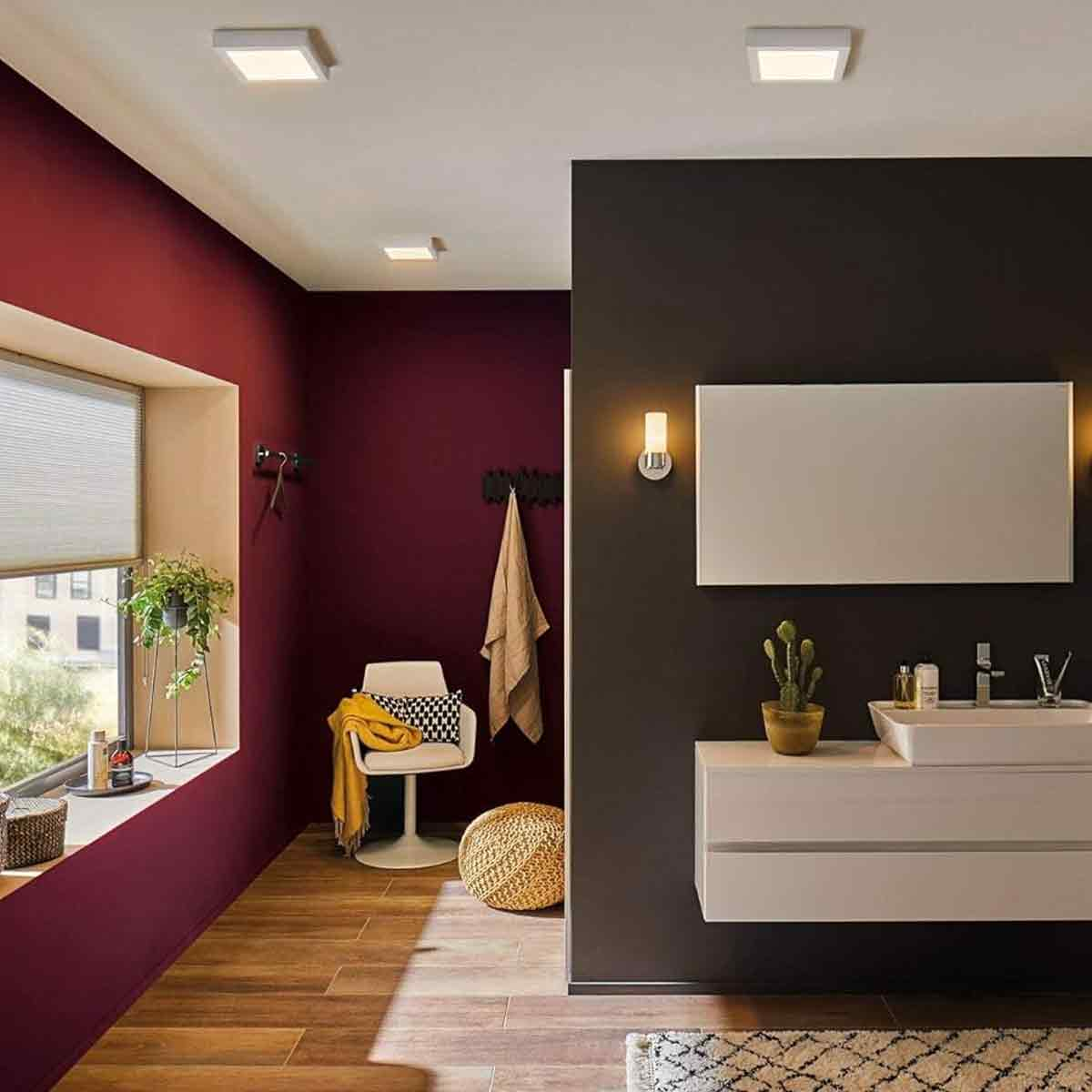 Ein modernes Badezimmer mit roten und schwarzen Wänden, Holzboden, einem weißen Waschtisch, einem Stuhl und einem Fenster mit Pflanzen.