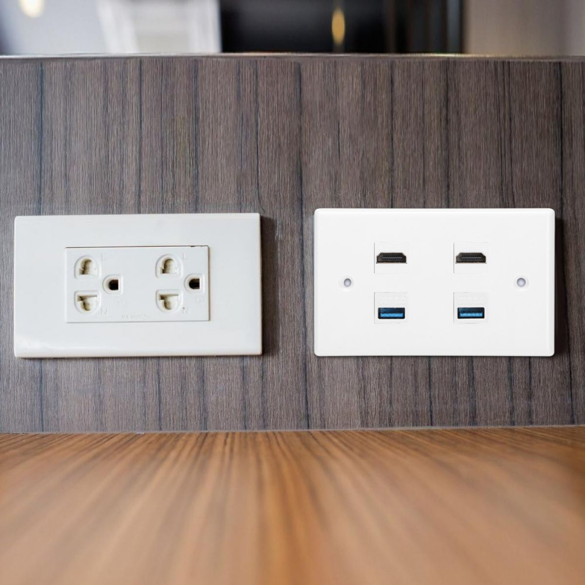 Eine Wand mit zwei weißen Paneelen. Eines hat eine Steckdose, das andere HDMI- und USB-Anschlüsse. Holztisch im Vordergrund.