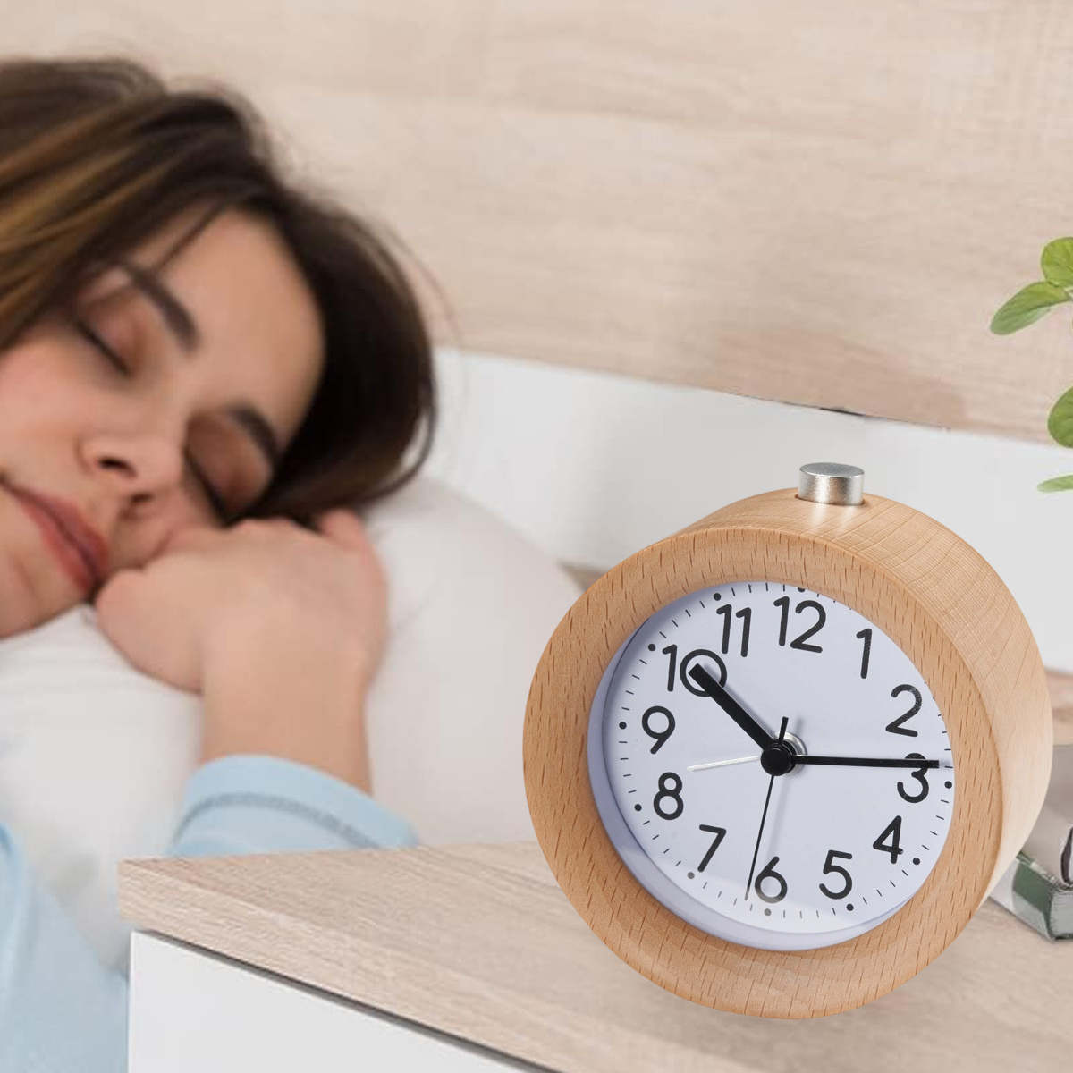 Eine Frau schläft im Bett neben einem Wecker aus Holz. Die Uhr zeigt 10:10. Im Hintergrund eine Pflanze.