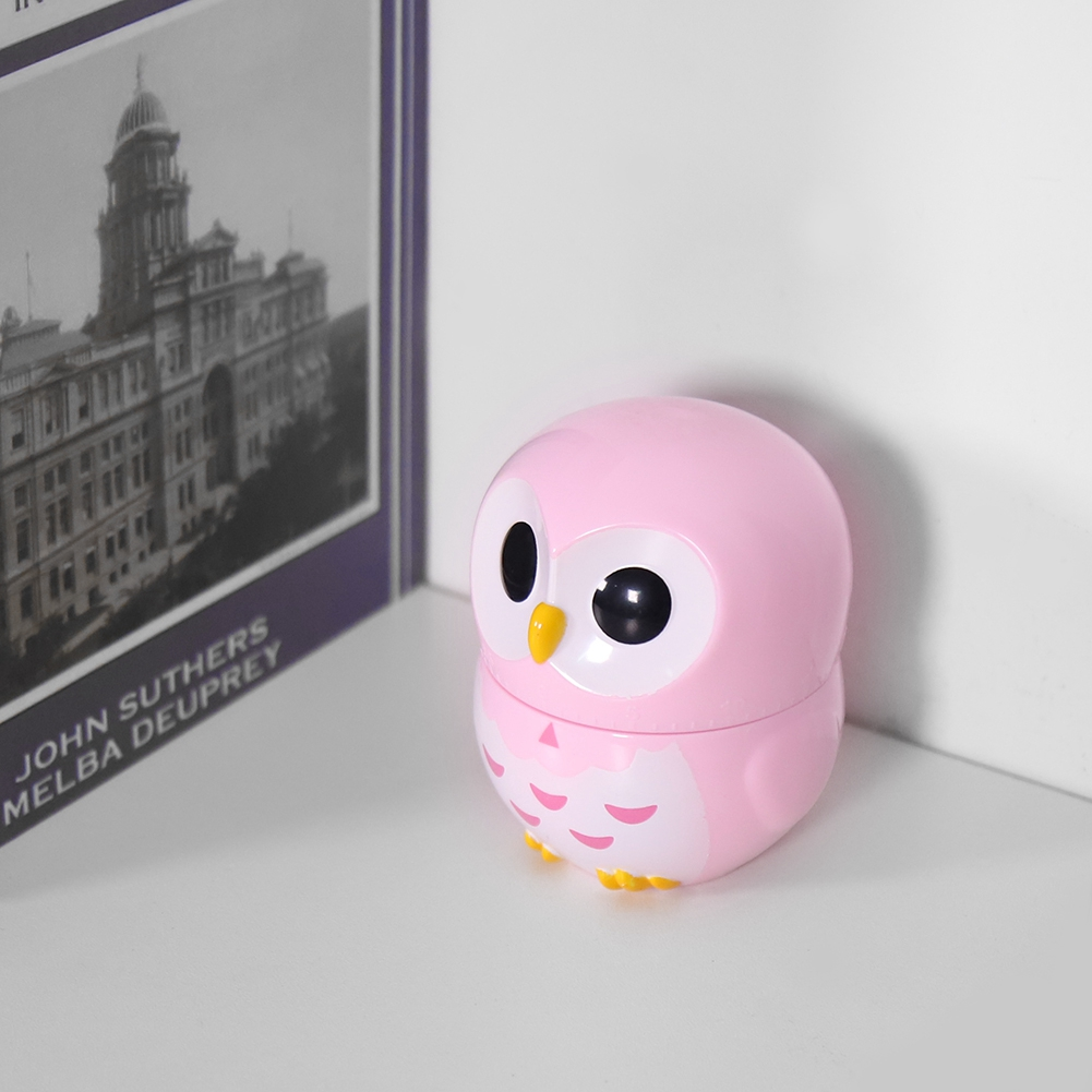 Ein rosa eulenformiger Behälter steht auf einer weißen Oberfläche. Er hat große schwarze Augen, einen gelben Schnabel und gelbe Füße. Im Hintergrund ist ein Buch.
