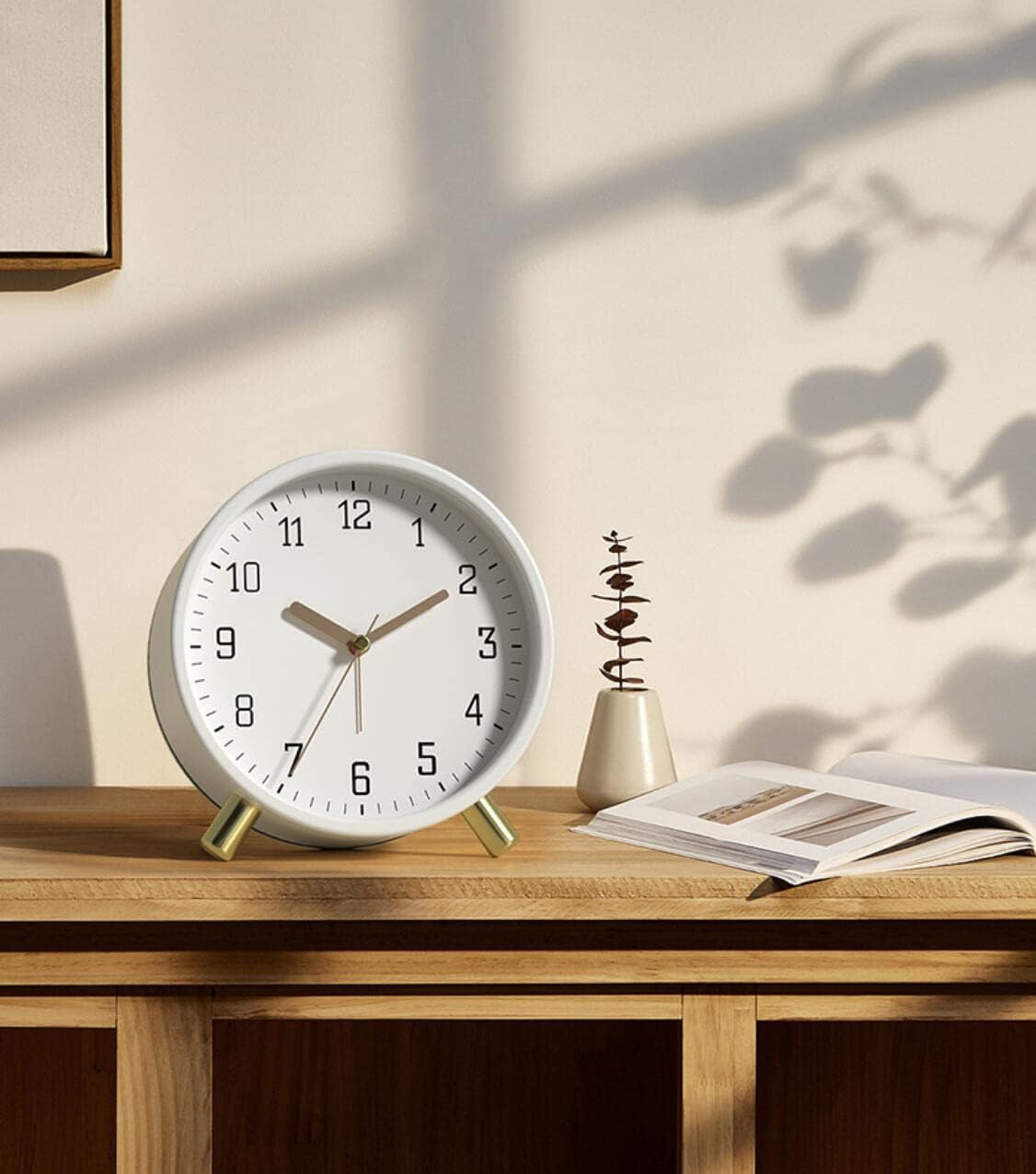Un orologio bianco con gambe dorate su una superficie di legno. Un vaso con una pianta e un libro aperto accanto. Ombre sul muro.