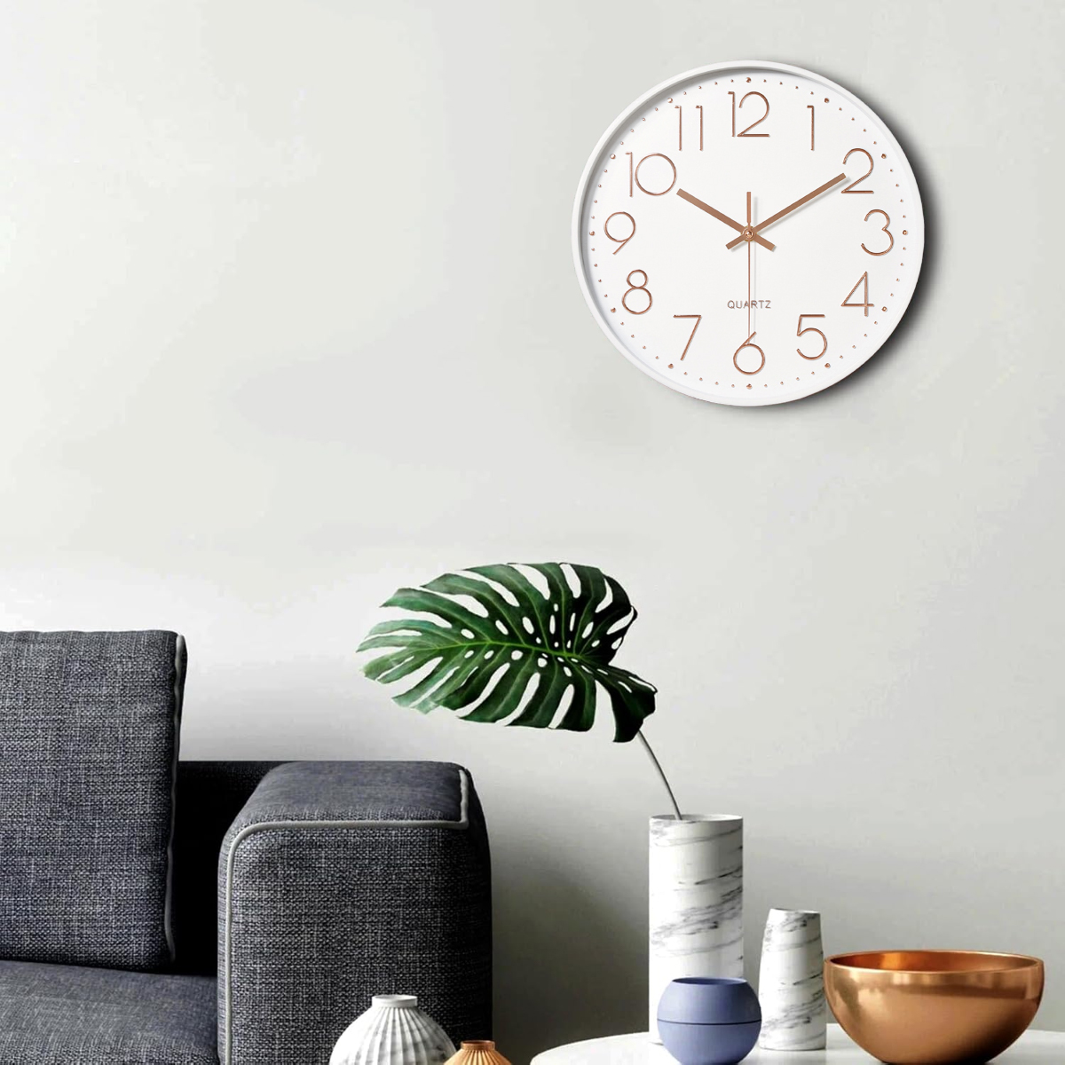 Weiße Uhr an der Wand, ein großes grünes Blatt in einer Vase und ein Sofa. Weitere Vasen und eine goldene Schale stehen auf einem Tisch.