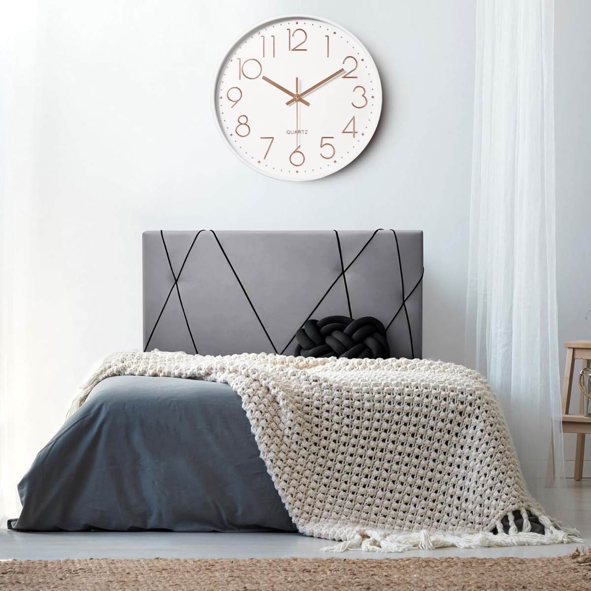 Schlafzimmer mit Bett, Uhr, grauem Kopfteil und beiger Decke. Das Bett ist mit einem grauen Laken bedeckt. Auf dem Bett liegt ein schwarzes Kissen.