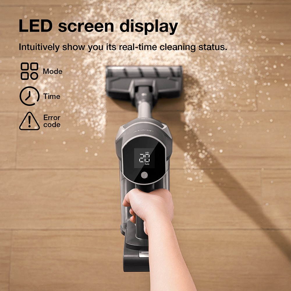 Mano che tiene un aspirapolvere con schermo LED. Lo schermo mostra l'ora e la modalità. Lo sfondo ha un pavimento in legno e polvere.