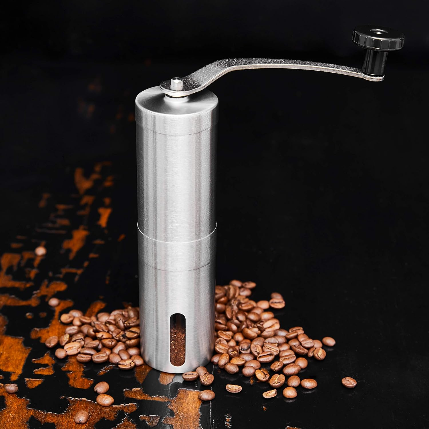 Ein silberner manueller Kaffeemühle steht auf einer dunklen Oberfläche mit verstreuten Kaffeebohnen. Die Mühle hat einen Griff.