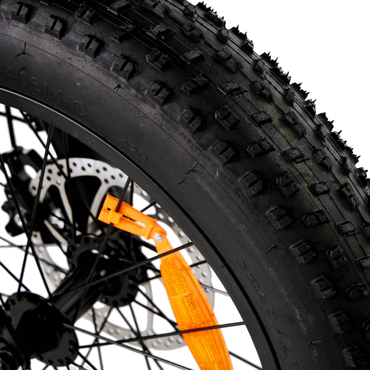 Zbliżenie czarnej opony rowerowej z głębokim bieżnikiem. Dołączony jest pomarańczowy odblask. Koło ma srebrne szprychy i hamulec tarczowy.