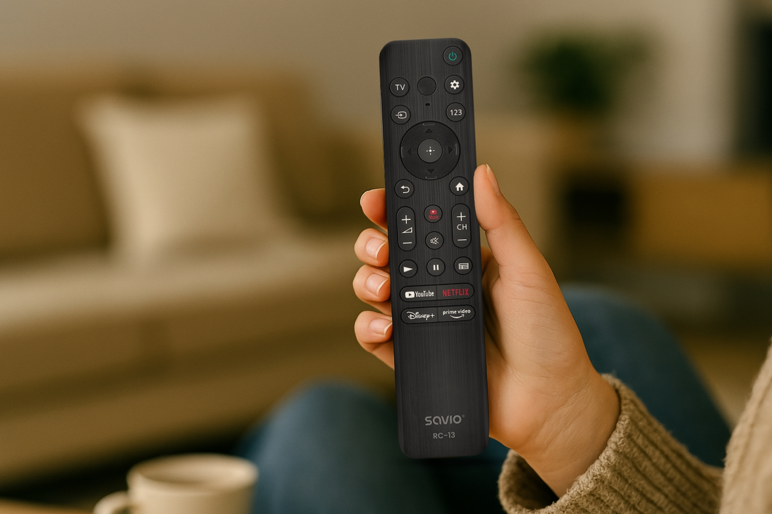 Dłoń trzyma czarny pilot z wieloma przyciskami. W tle rozmyty widok salonu.