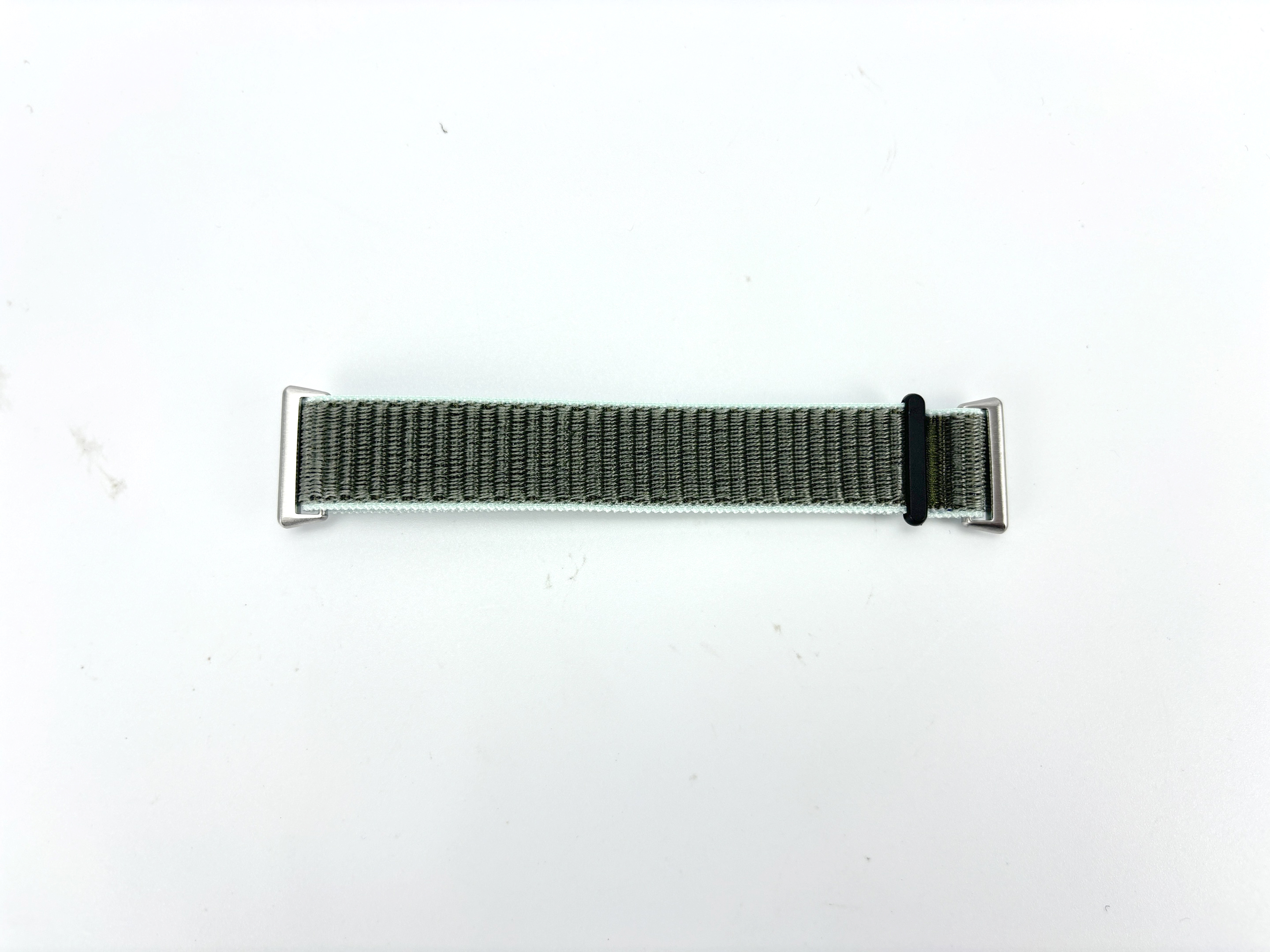 Ein grünes und graues Uhrenarmband liegt auf einer weißen Oberfläche. Es hat silberne Enden und eine schwarze Schlaufe.