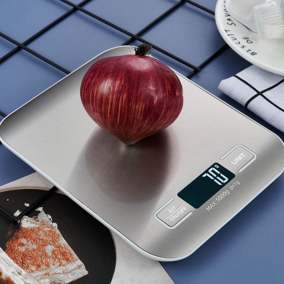 Eine rote Zwiebel auf einer silbernen Waage mit Digitalanzeige von 70g. Im Hintergrund ein Teller und eine Serviette.