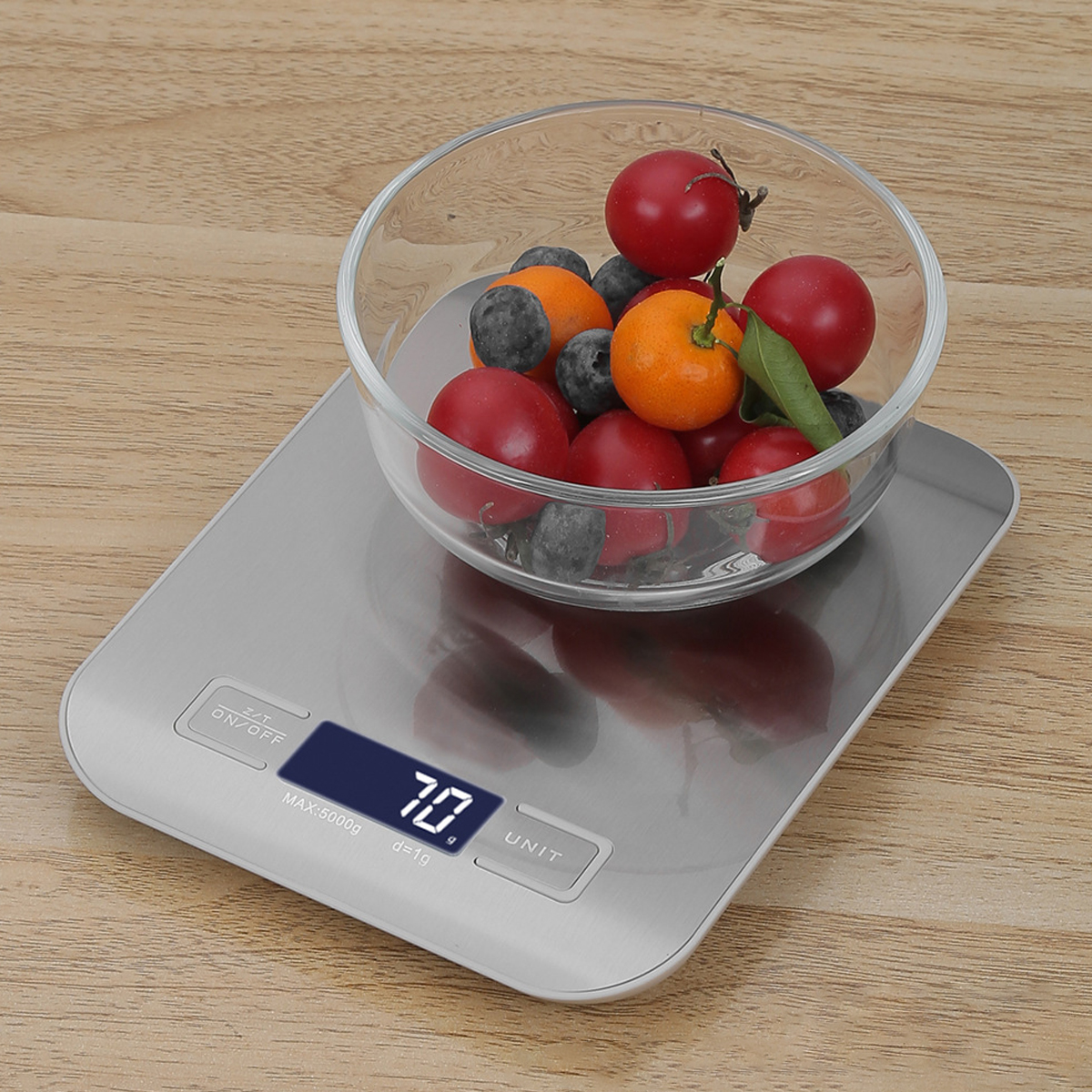 Eine Glasschüssel mit Tomaten, Blaubeeren und einer orangefarbenen Frucht steht auf einer digitalen Küchenwaage, die 70 anzeigt.