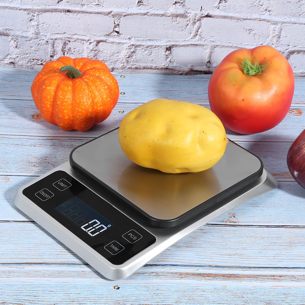 Eine digitale Küchenwaage mit einer Kartoffel darauf. Ein Kürbis und eine Tomate sind in der Nähe. Die Waage zeigt 0,0 g an.