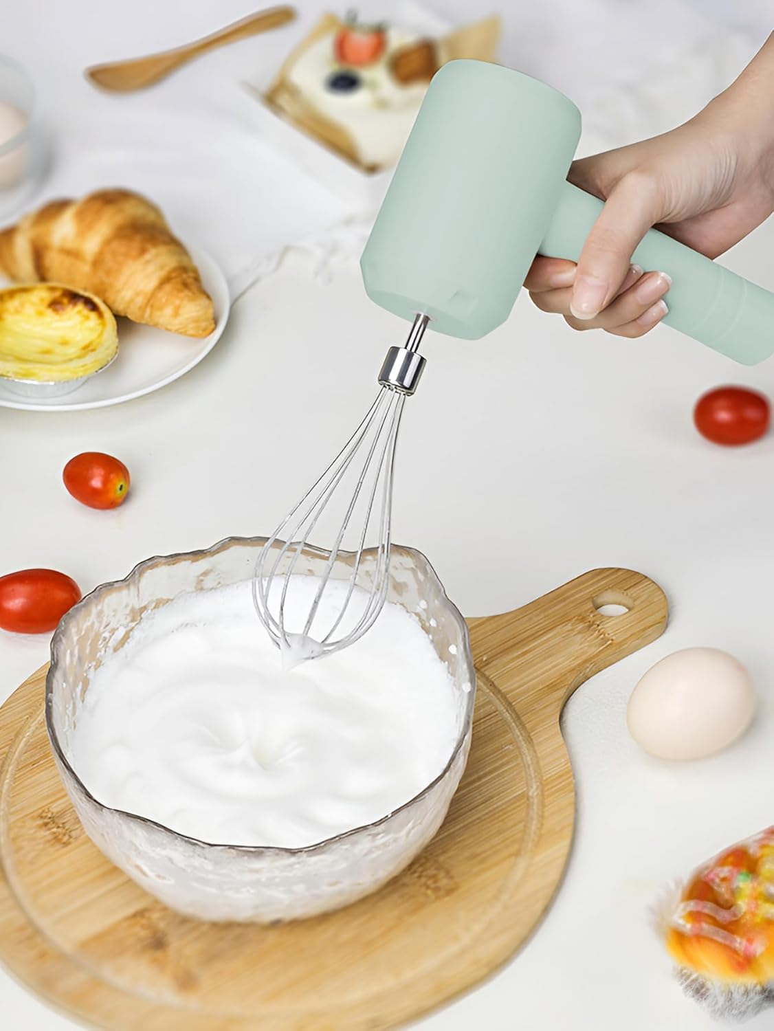Eine Hand hält einen türkisfarbenen Mixer über einer Schüssel mit geschlagenem Eiweiß. Weitere Artikel sind Tomaten, Gebäck und ein Ei.