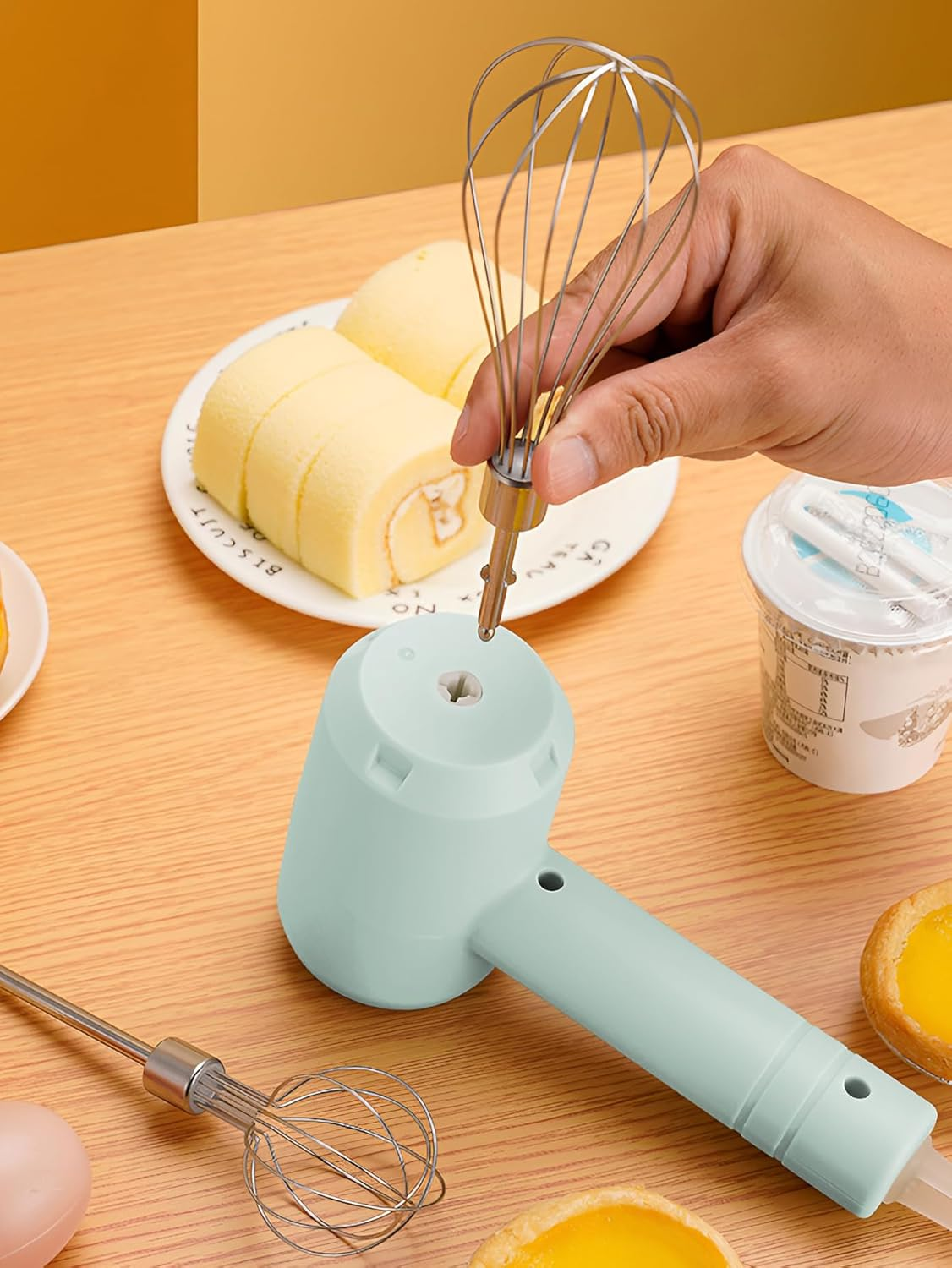 Eine Hand hält einen Schneebesen und steckt ihn in einen hellblauen elektrischen Mixer. Auf dem Tisch liegen Kuchenrollen, Eiertörtchen und Joghurt.