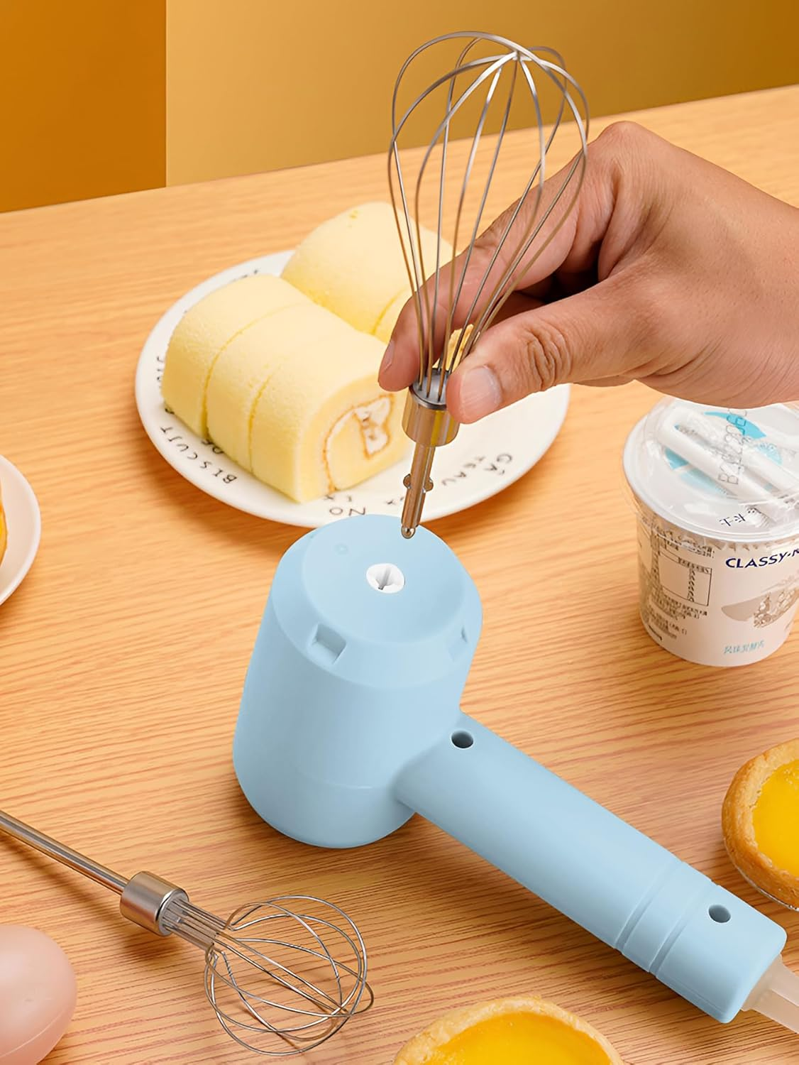 Eine Hand steckt einen Schneebesen in einen blauen Elektromixer. Kuchenrollen und ein Joghurtbecher liegen auf dem Holztisch. Auch ein Kuchen ist vorhanden.