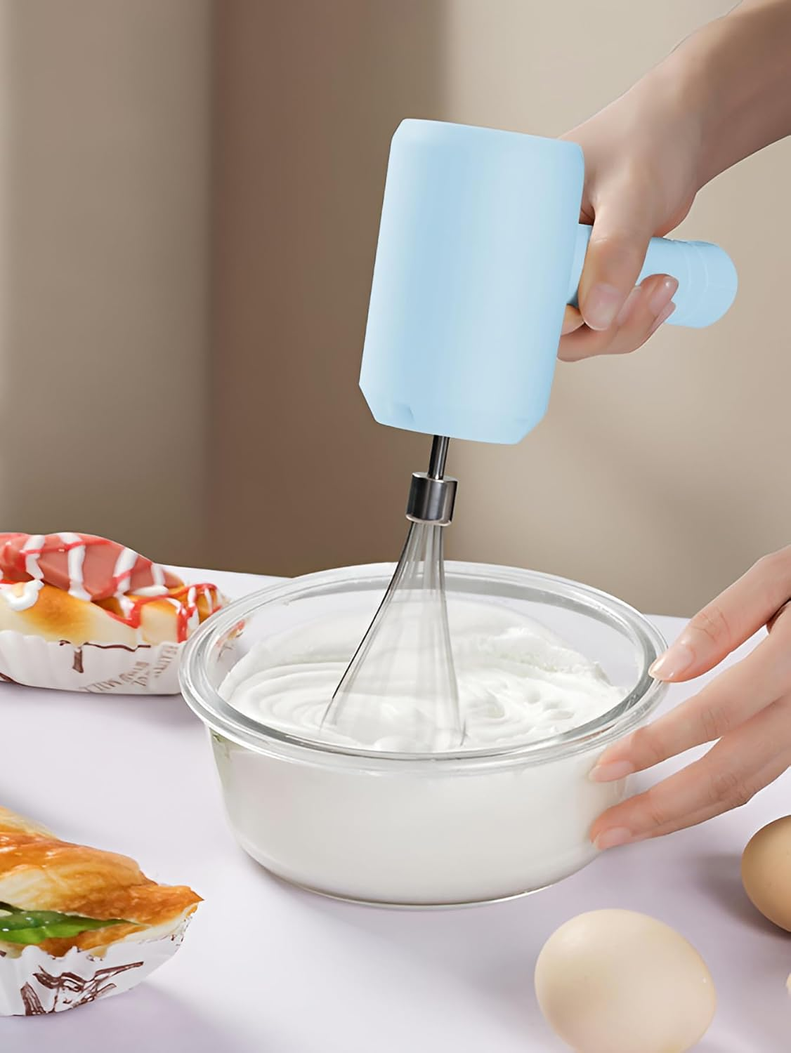 Eine Person verwendet einen blauen Handmixer, um Sahne in einer Glasschüssel zu schlagen. Eier und Gebäck liegen auf dem Tisch.