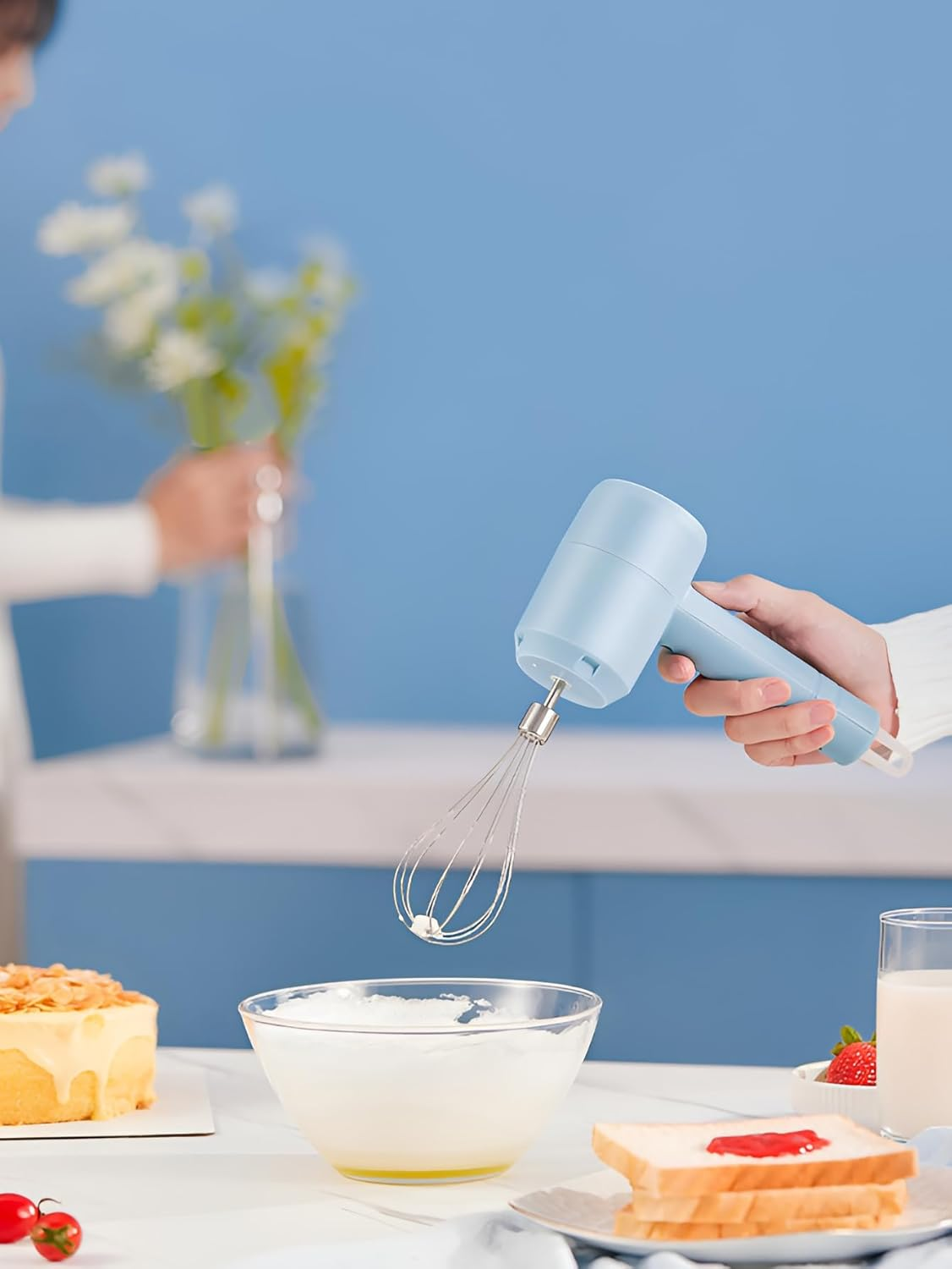 Eine Person verwendet einen blauen Handmixer, um Sahne zu schlagen. Auf dem Tisch befinden sich Kuchen, Brot mit Marmelade und andere Lebensmittel.