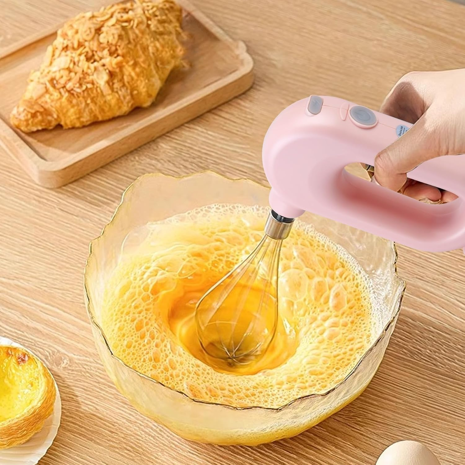 Ein Handrührgerät verrührt Eier in einer Glasschüssel. Ein Croissant und ein Gebäck liegen auf dem Holztisch.